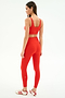 A woman, facing away and looking to the side, wears SPLITS59 Airweight Rib High Waist 7/8 leggings in Lotus Red with a matching sports bra and white Adidas sneakers against a plain white background.