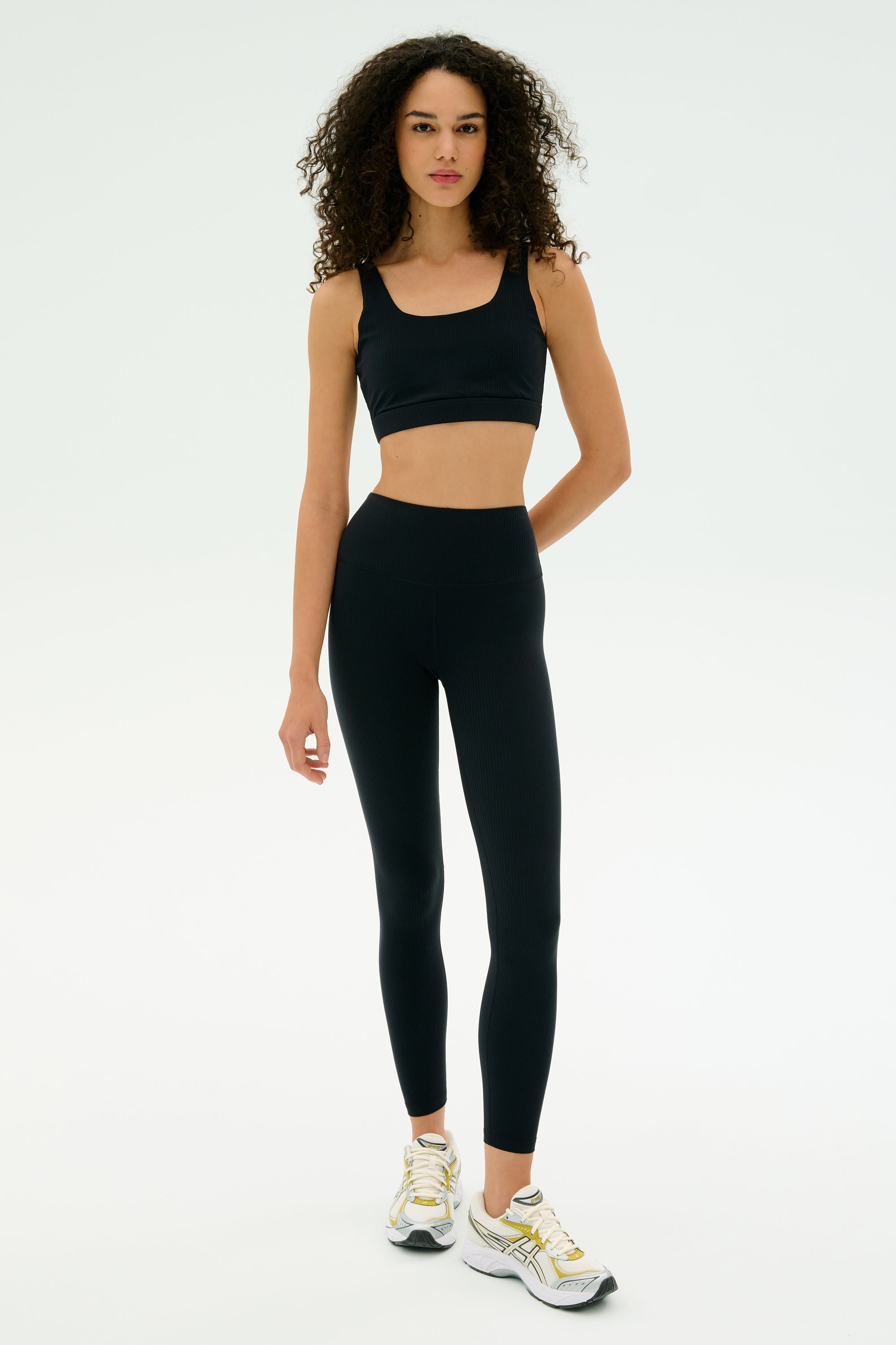 A woman wears the SPLITS59 Airweight Rib Bra in black, paired with black high-waisted leggings and white athletic shoes against a plain white background.