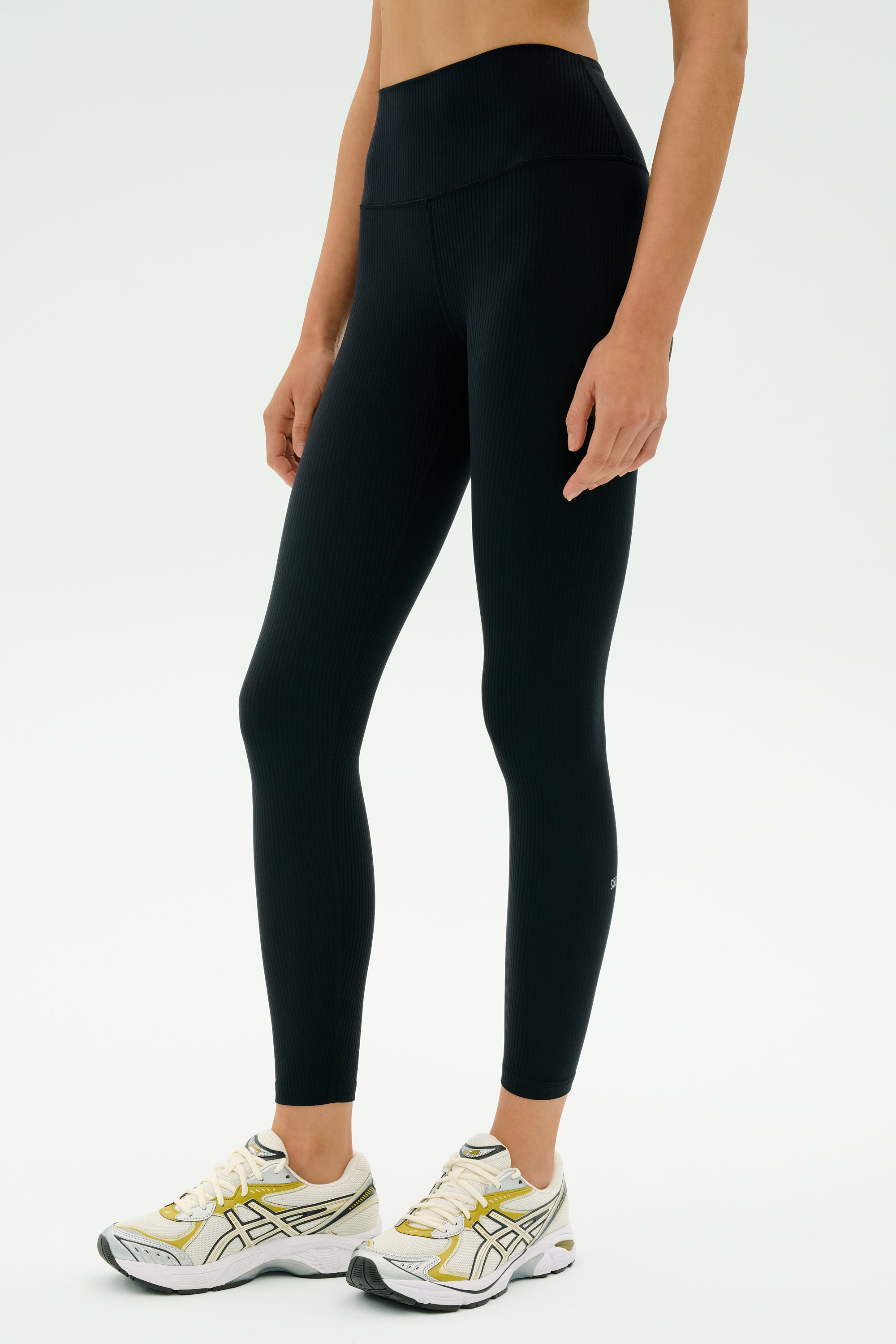 A person wearing SPLITS59 Airweight Rib High Waist 7/8 black leggings and white athletic shoes stands against a plain light background.