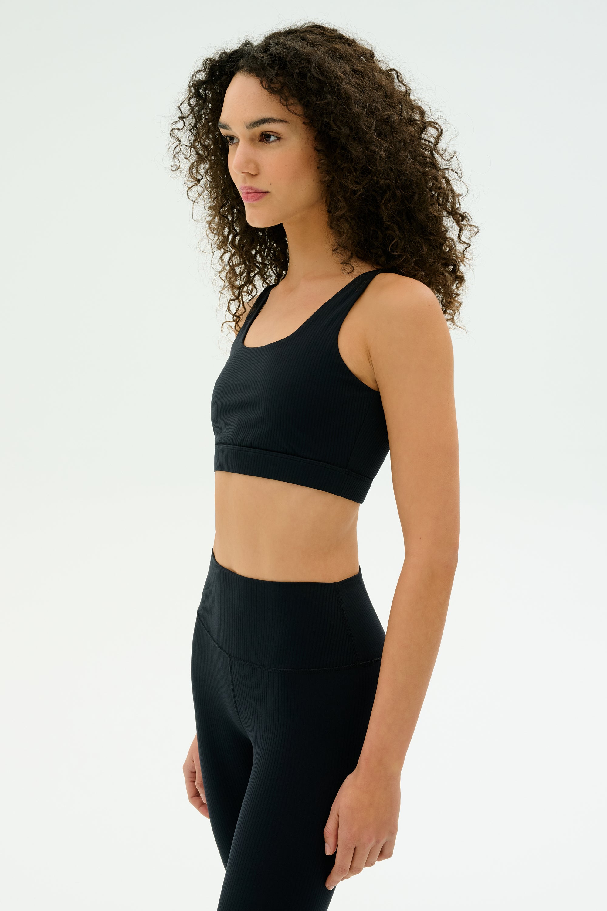 A woman with curly hair models the SPLITS59 Airweight Rib Bra in black, paired with matching leggings, standing against a plain white background and facing slightly to the side.