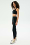A woman with curly hair stands in profile, wearing the SPLITS59 Airweight Rib Bra in black, paired with matching leggings and white athletic shoes, all set against a plain white background.