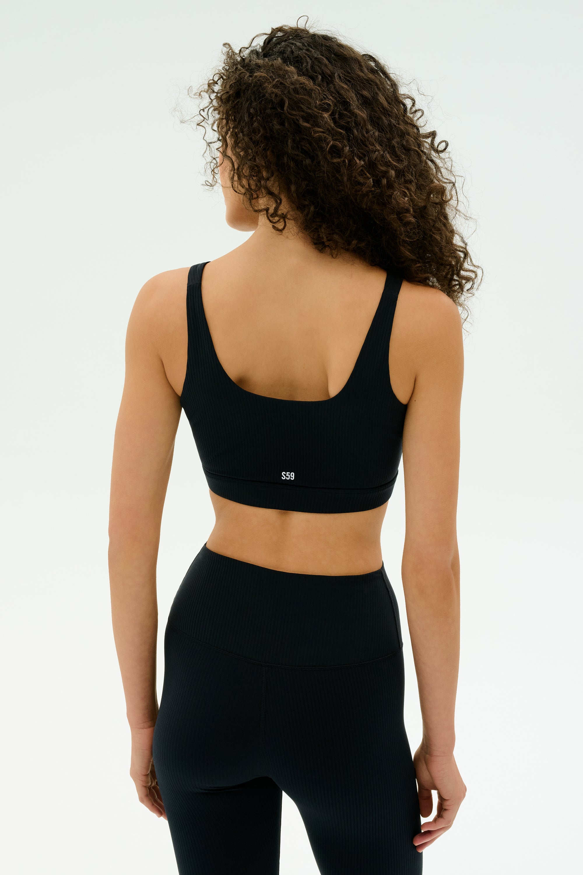A person with curly hair faces away against a plain light background, wearing the SPLITS59 Airweight Rib Bra in black and matching leggings.