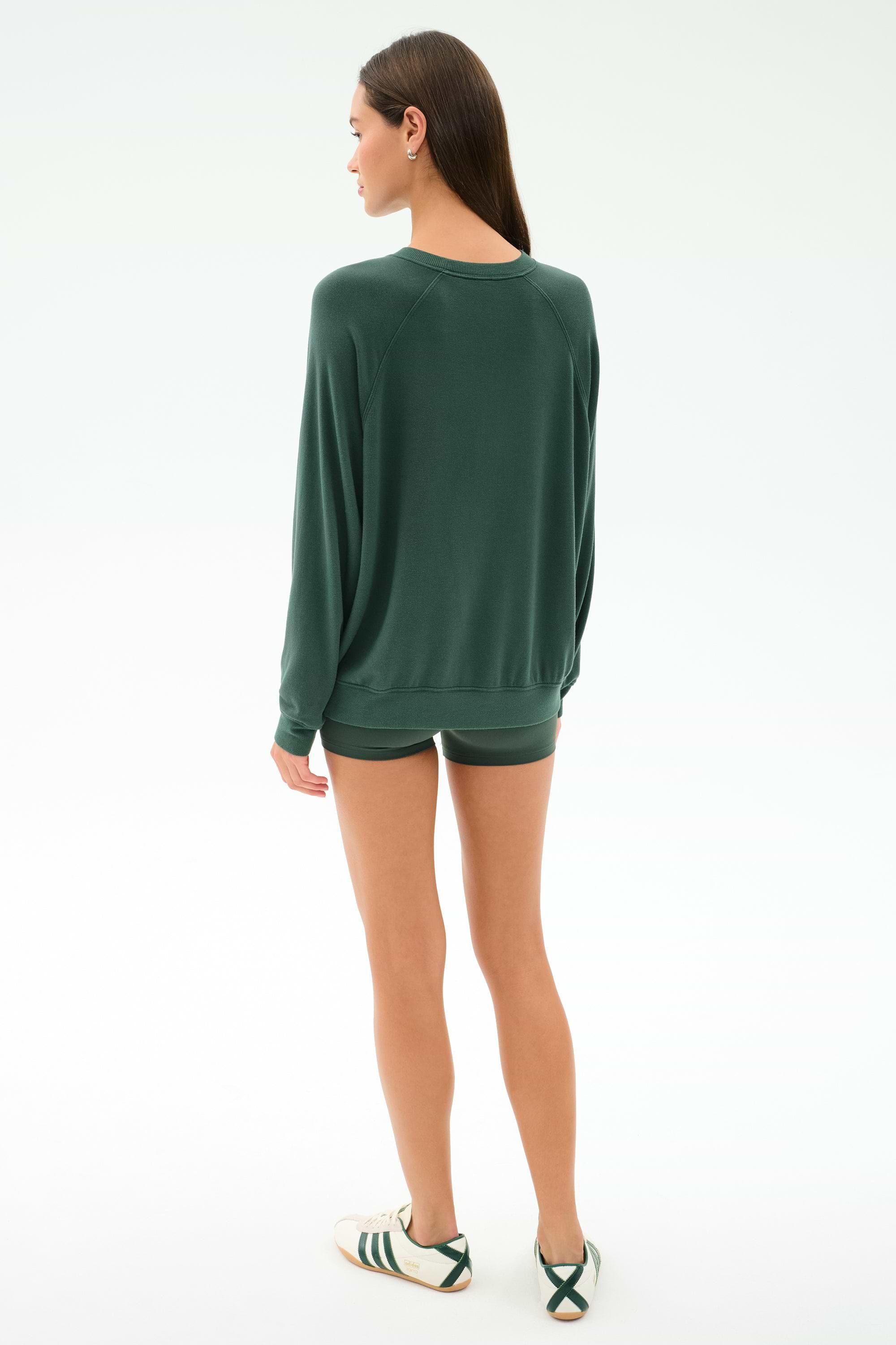 A woman stands facing away in a SPLITS59 Andie Fleece Sweatshirt - Military, matching green shorts, and white sneakers with green stripes against a plain white background.