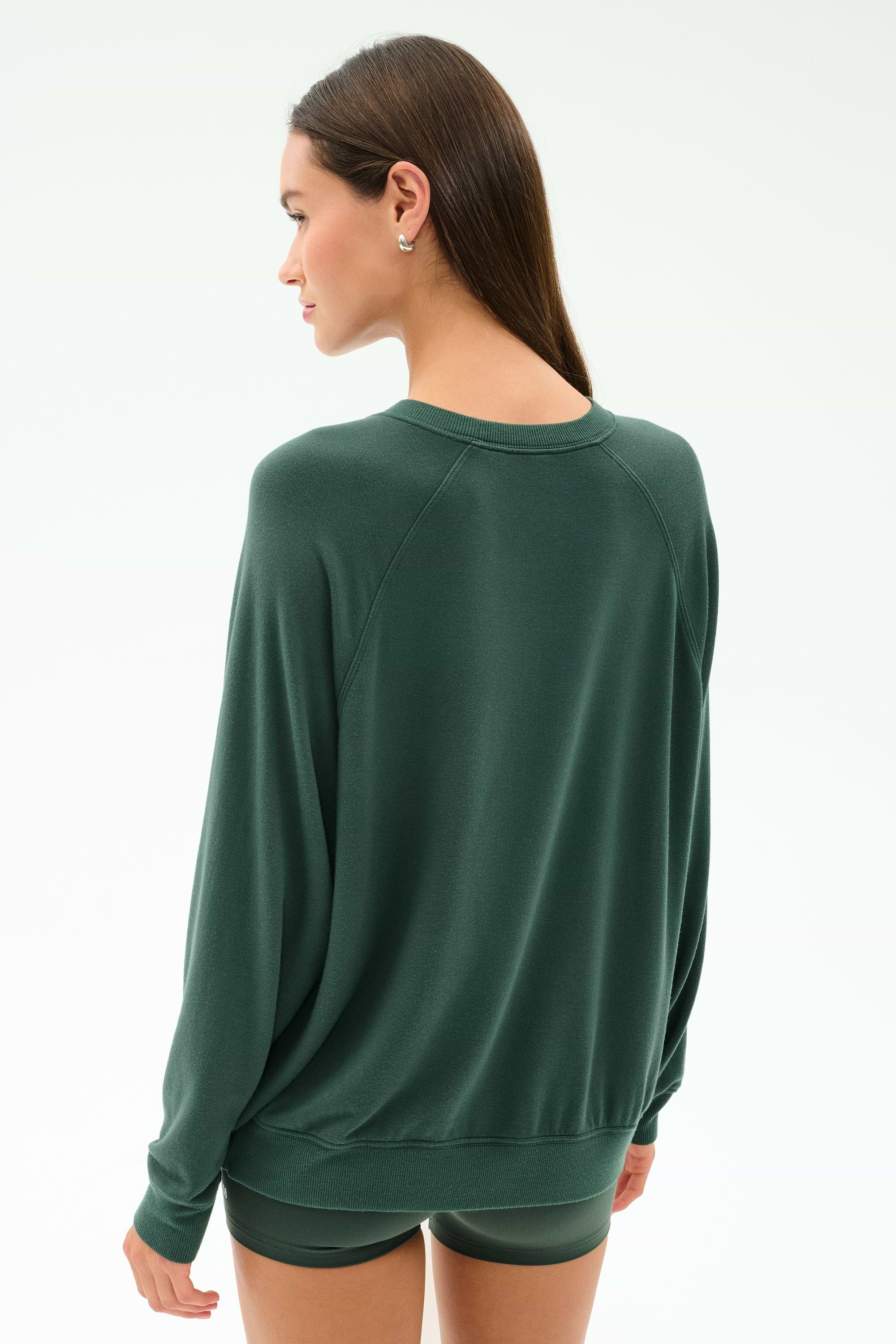 A person with long brown hair stands facing away from the camera, wearing the SPLITS59 Andie Fleece Sweatshirt in Military, a dark green pullover made in Los Angeles.