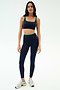 A woman faces forward in SPLITS59’s Easton Rigor High Waist 7/8 leggings and matching indigo/grass sports bra, made of compressive fabric, paired with white sneakers against a plain white background.