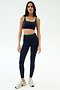 A woman faces forward in SPLITS59’s Easton Rigor High Waist 7/8 leggings and matching indigo/grass sports bra, made of compressive fabric, paired with white sneakers against a plain white background.