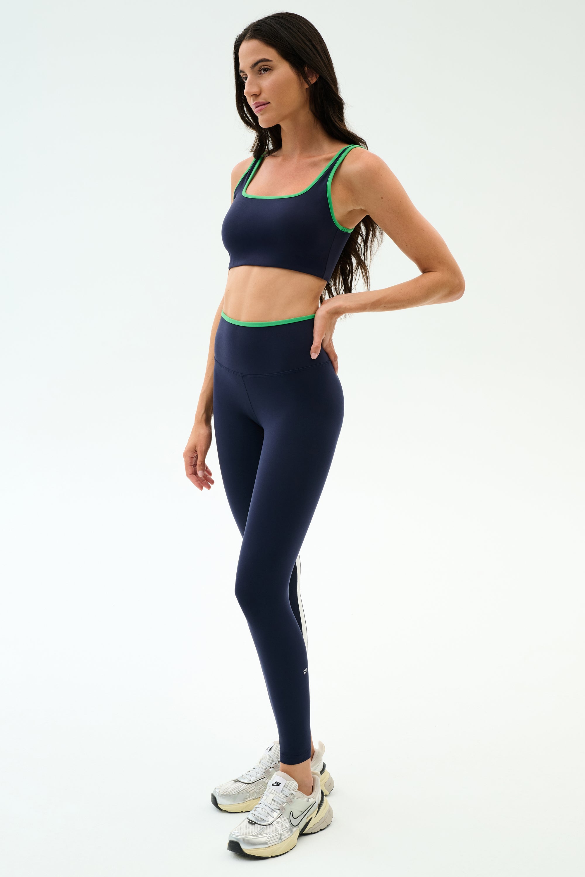 A woman stands against a white background wearing the SPLITS59 Cait Rigor Bra in Indigo/Grass and matching navy leggings, both crafted from Rigor fabric—ideal for high-impact workouts.