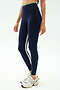 A person wears SPLITS59 Easton Rigor High Waist 7/8 leggings in Indigo/Grass—navy blue with side stripes, made from compressive fabric—with white sneakers, shown from shoulders down against a plain background.