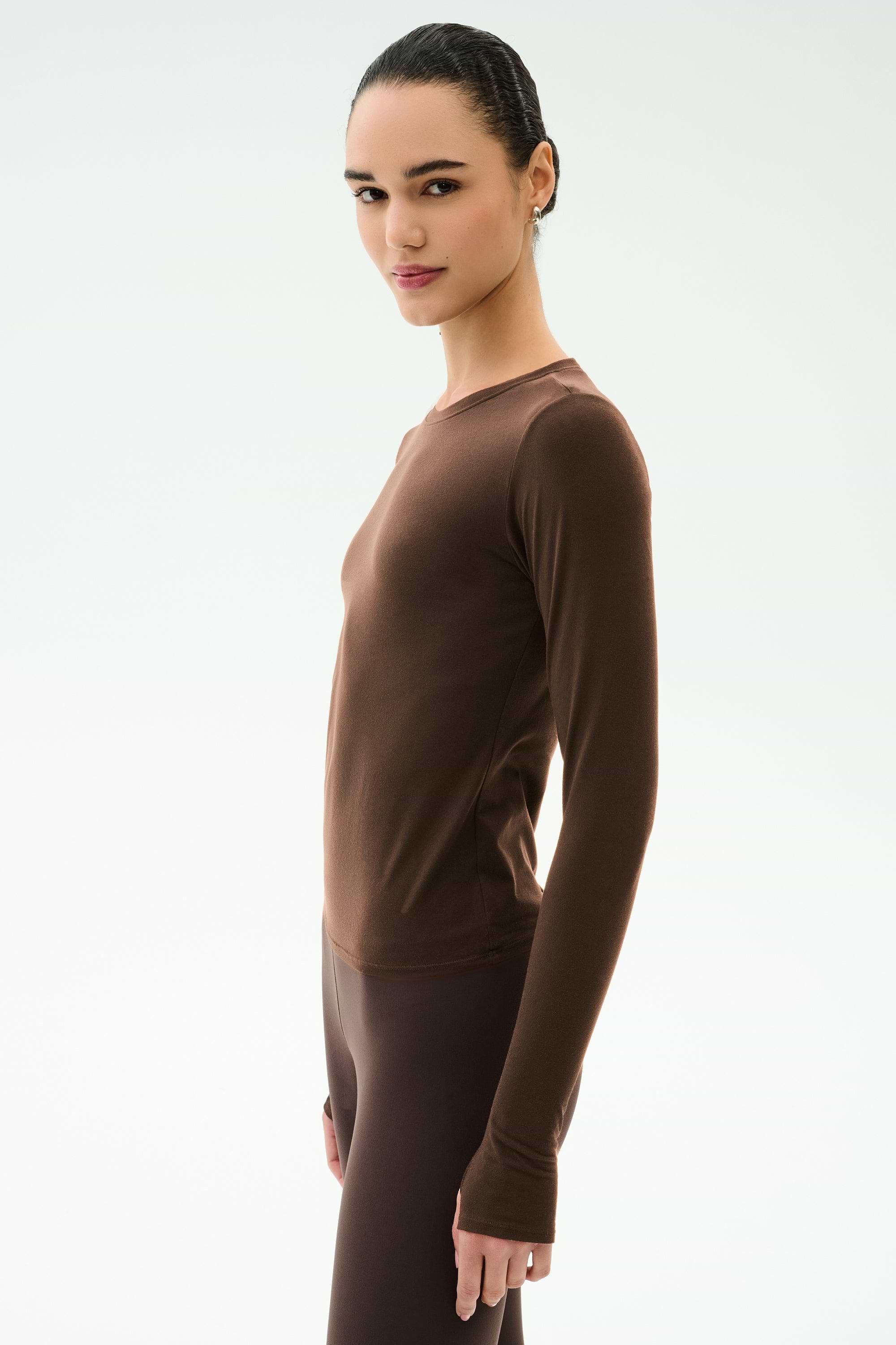 A person with dark hair tied back, wearing the SPLITS59 Airweight Lite Long Sleeve Waist Length top in Dark Chocolate and matching leggings, stands in profile against a plain light background.