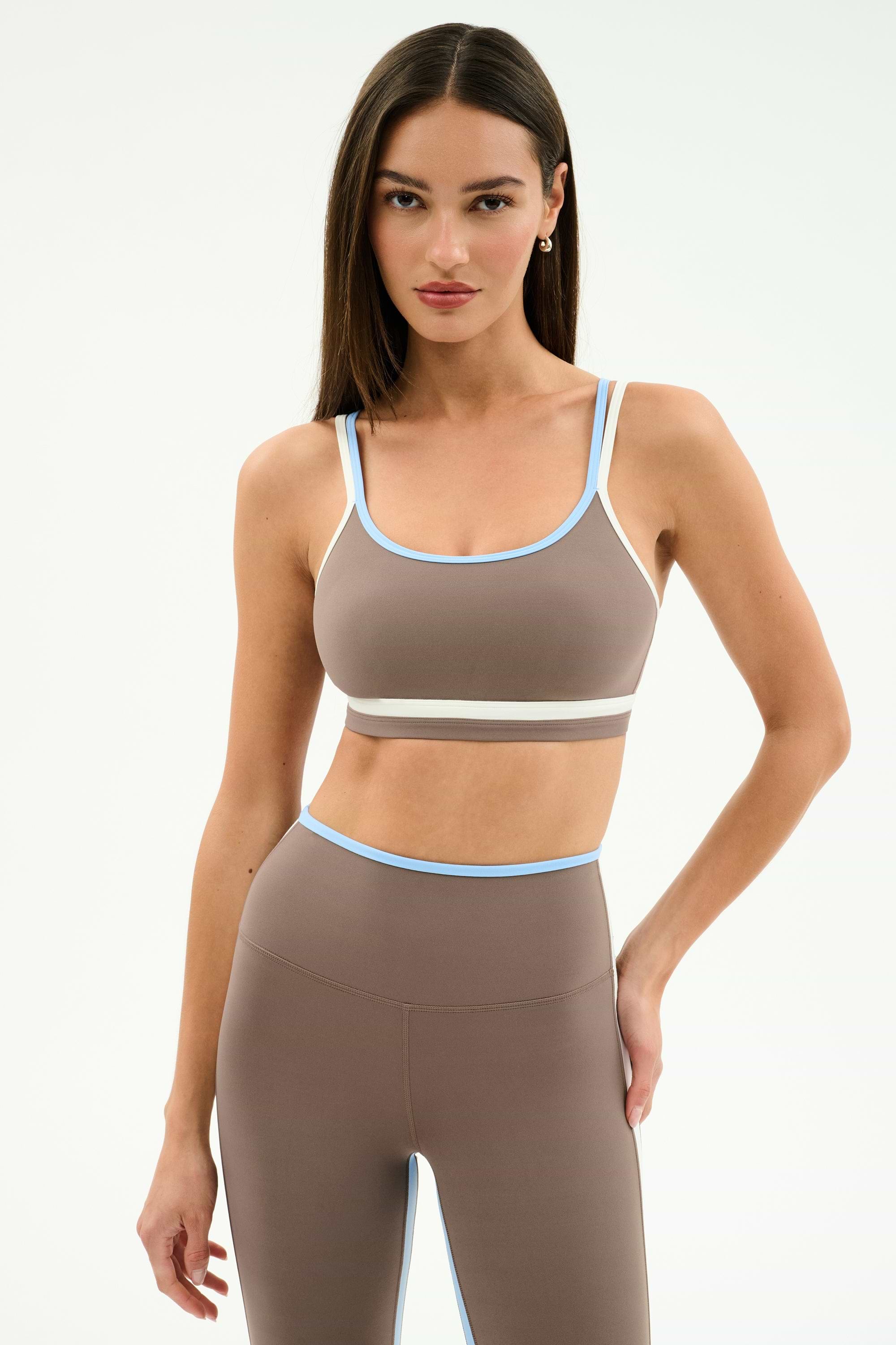 A woman stands against a plain background wearing the SPLITS59 Eva Rigor Double Strap Bra in Lentil/White, perfect for yoga or workouts, paired with matching high-waisted leggings.
