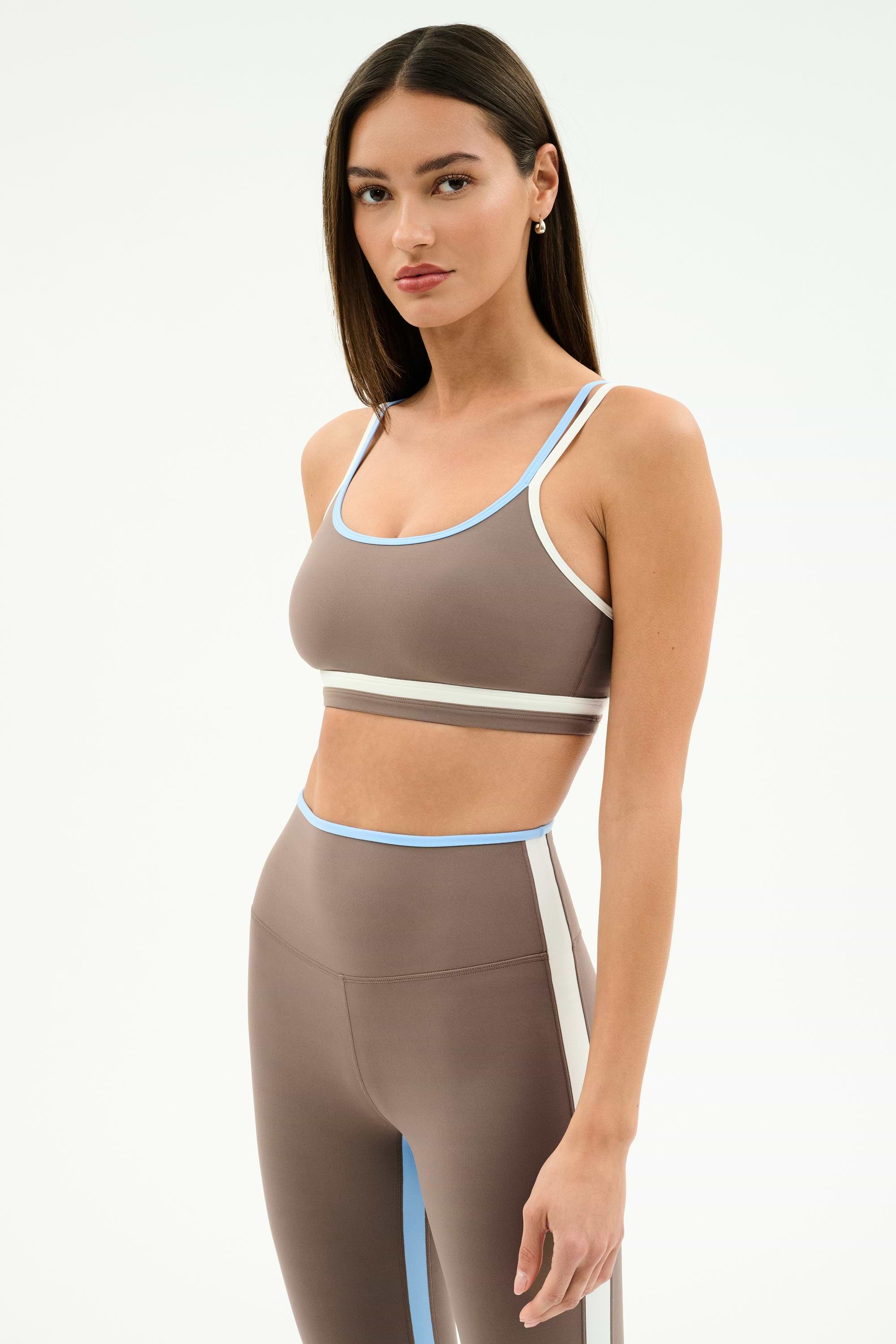 A woman stands against a plain light background, wearing the SPLITS59 Eva Rigor Double Strap Bra in Lentil/White with matching high-waisted leggings featuring light blue and white trim.