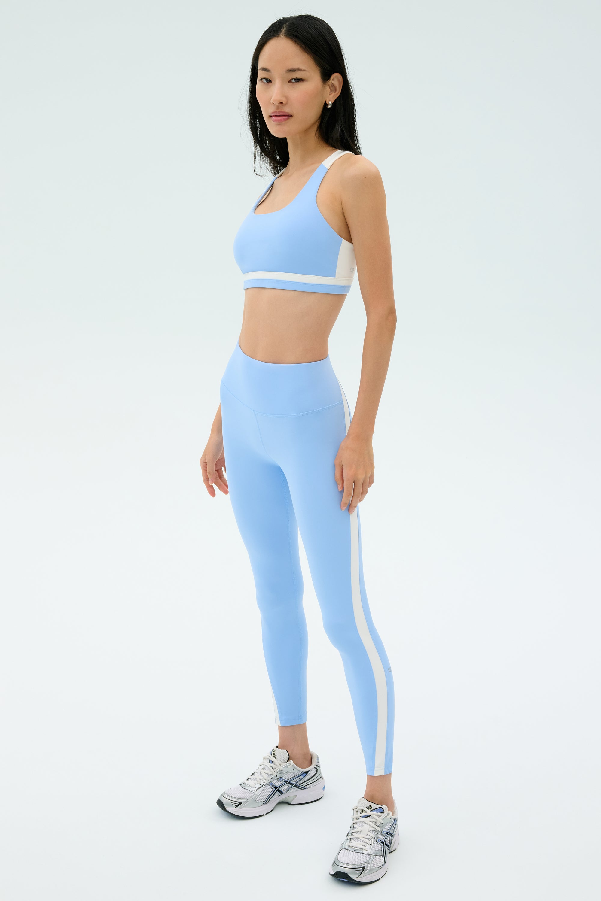 A woman wears the SPLITS59 Miles Rigor Bra in Skylight/White with matching leggings and silver athletic shoes, standing against a plain light background.