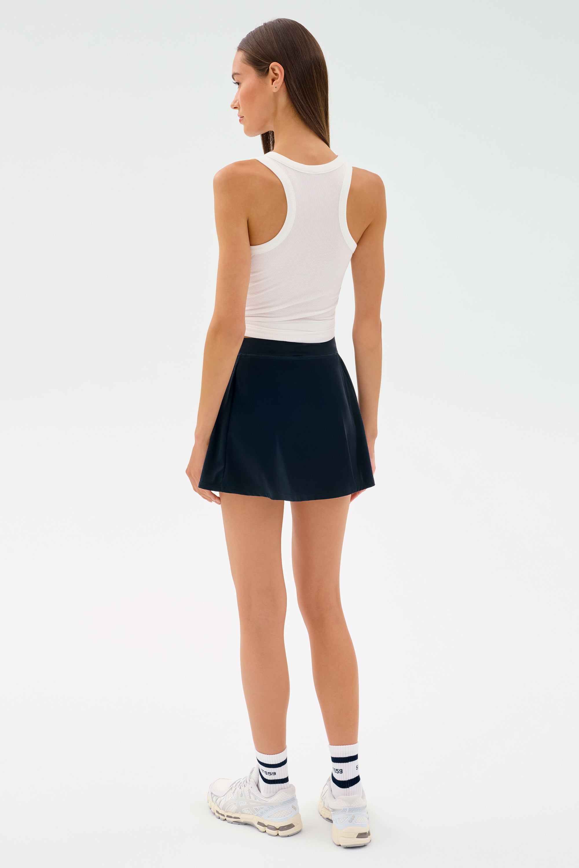 A woman faces away against a plain light background, wearing a white tank top, the SPLITS59 Venus High Waist Rigor Skort in Indigo, white sneakers, and striped white socks.