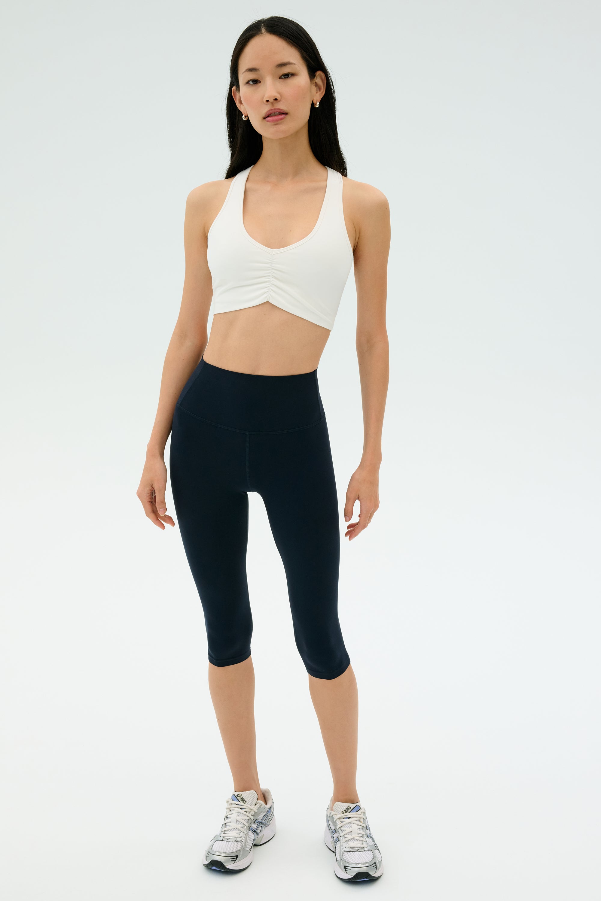 A woman stands facing forward against a plain light background, wearing the SPLITS59 Airweight Bra with Ruching in white, navy blue athletic leggings, and silver running shoes.