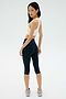 A woman stands on a white background in the SPLITS59 Airweight Bra with Ruching in white, paired with black high-waisted capri leggings and white athletic shoes—an ideal look for hot yoga—as she looks over her shoulder.