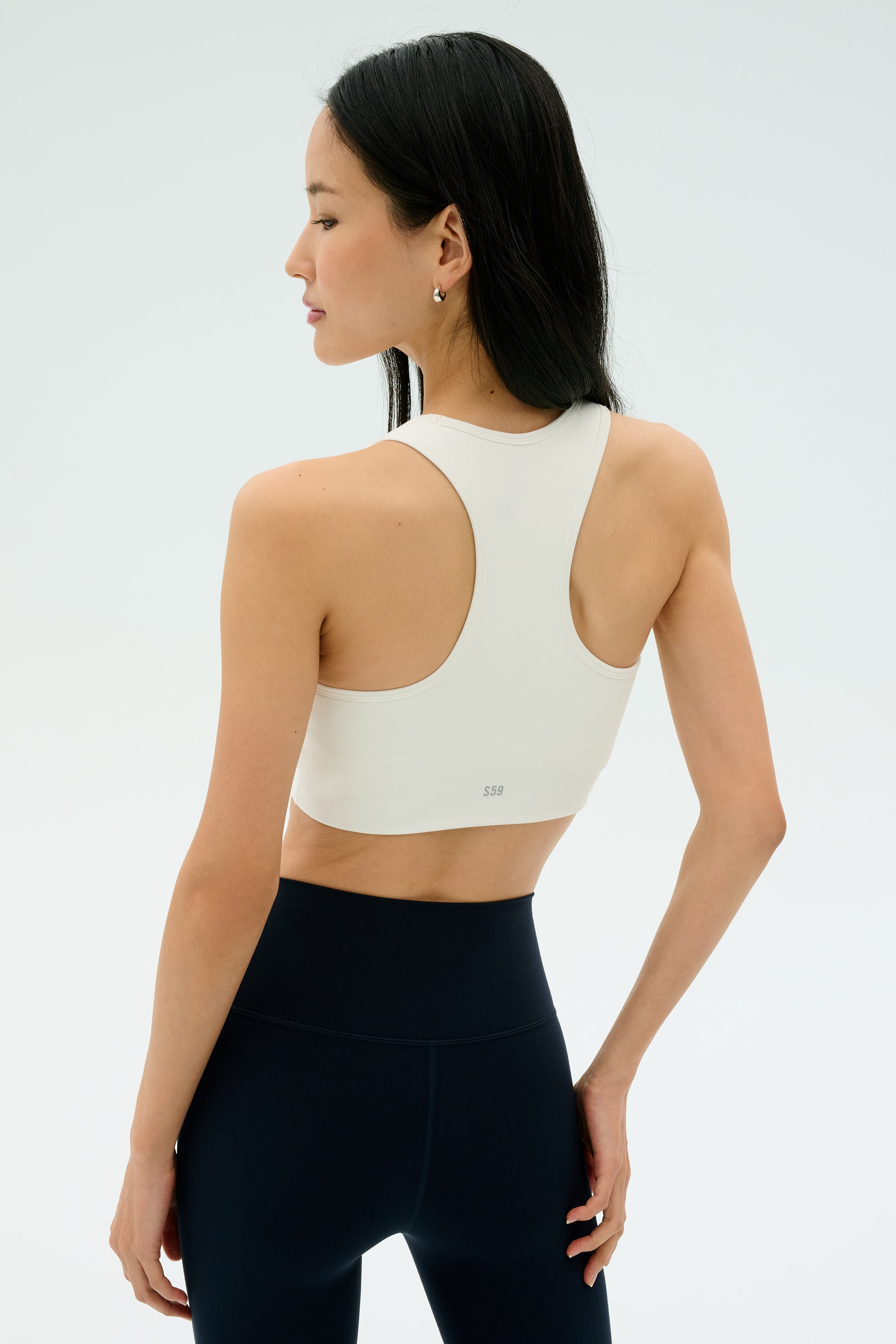A woman stands facing away against a plain light background, wearing the SPLITS59 Airweight Bra with Ruching in white paired with high-waisted black leggings.