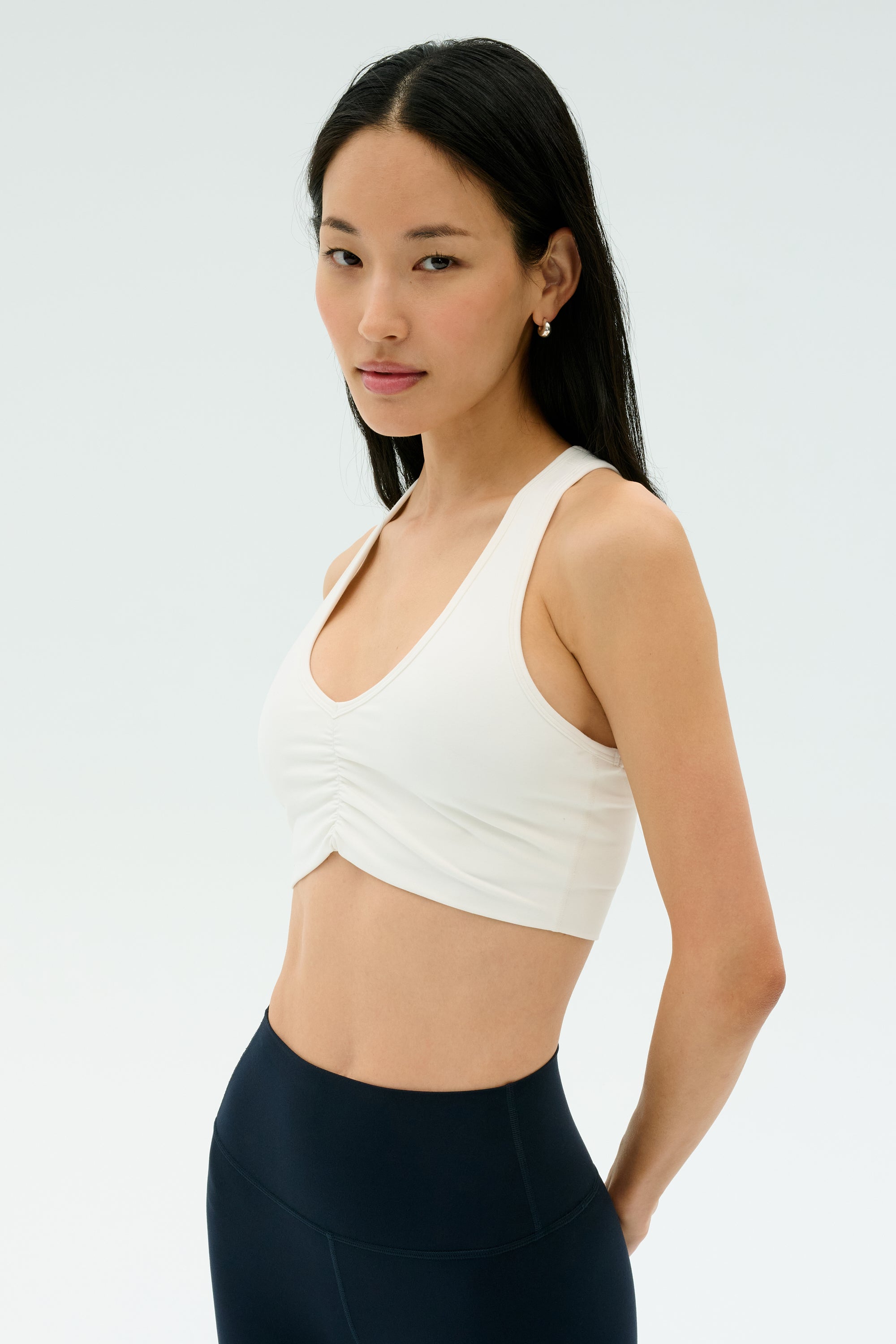 A woman with long dark hair models the SPLITS59 Airweight Bra with Ruching in white, paired with navy leggings, standing against a plain light background.
