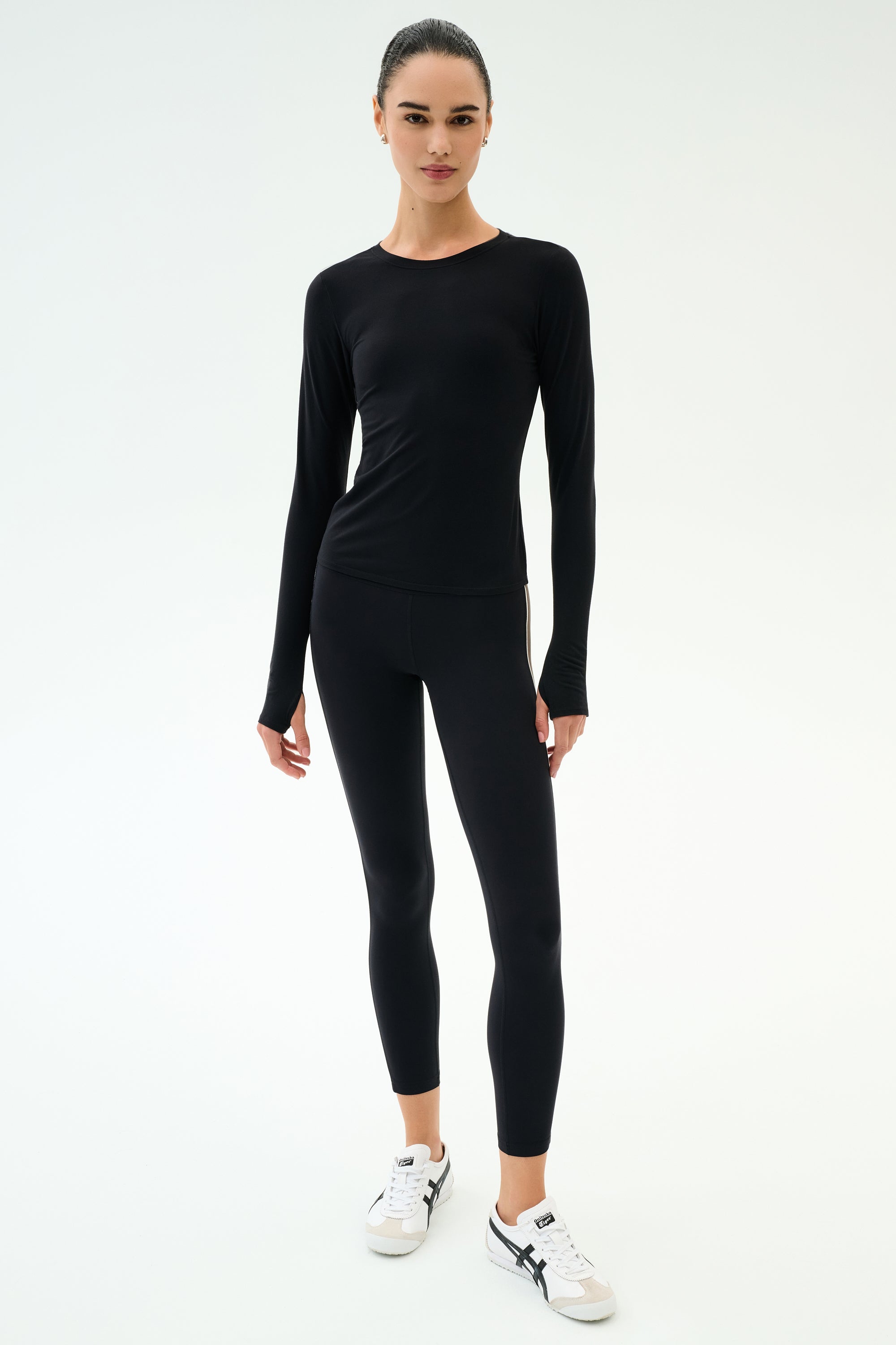 A person stands against a white background wearing the SPLITS59 Airweight Lite Long Sleeve Waist Length top in black, paired with black leggings and white sneakers—an ideal outfit for gym workouts.