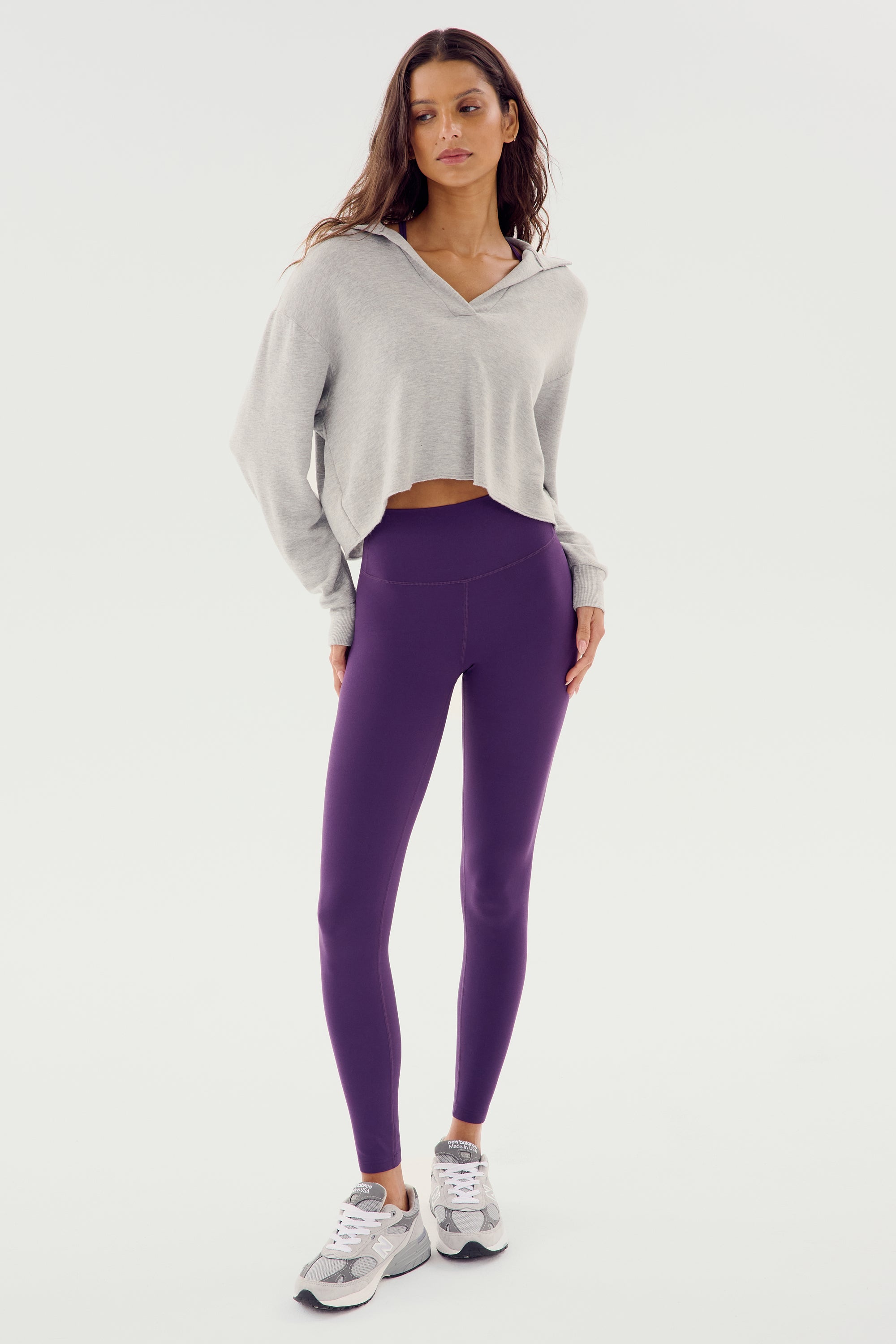 A woman is standing and wearing a heather grey Joan Fleece Polo Sweatshirt by SPLITS59, purple leggings, and white sneakers. She has long, wavy hair and is looking towards the camera. The background is plain and white.