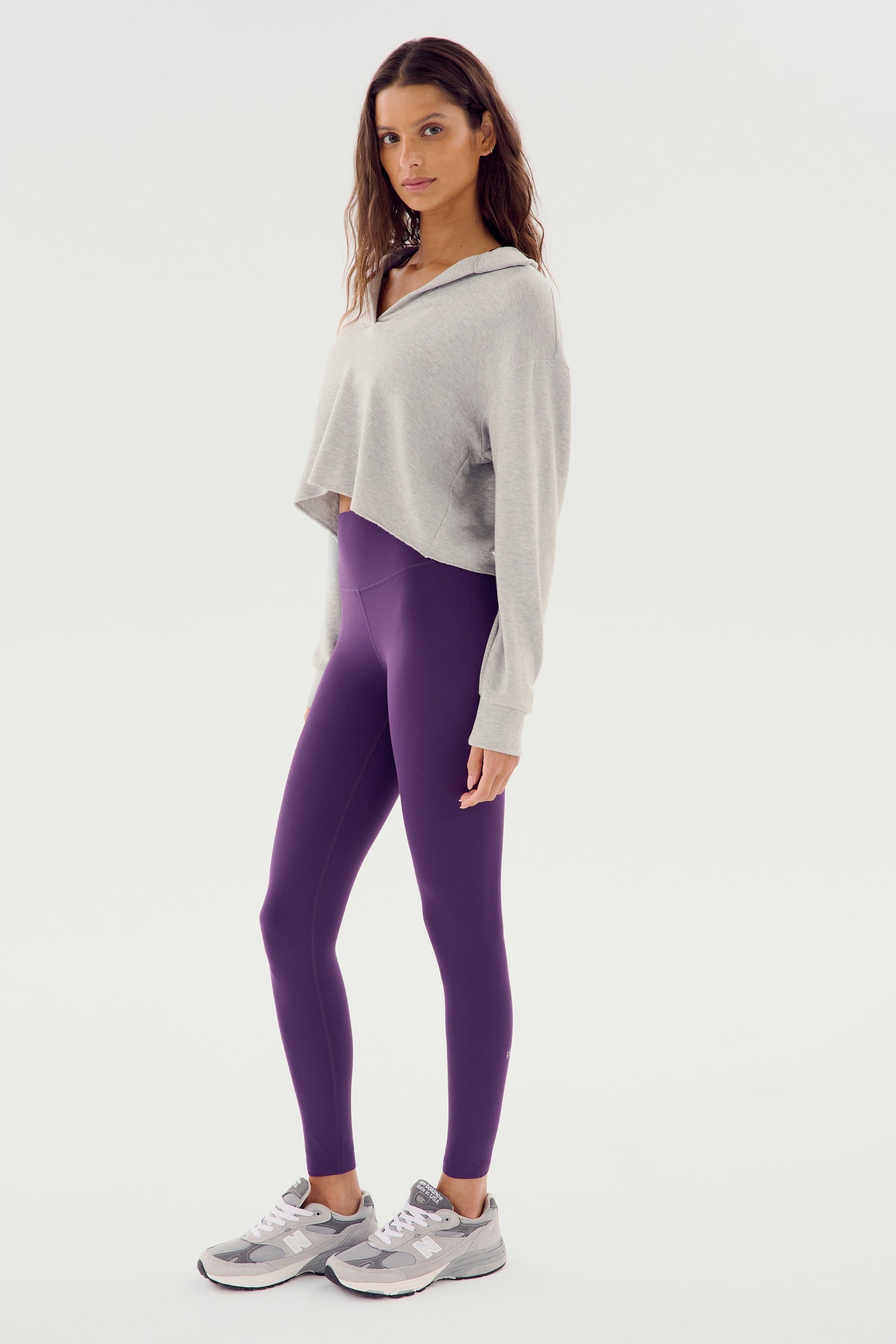 A woman stands in a relaxed pose wearing a Joan Fleece Polo Sweatshirt in Heather Grey by SPLITS59, along with purple leggings and grey sneakers made of 95% MODAL and 5% SPANDEX, against a plain white background.