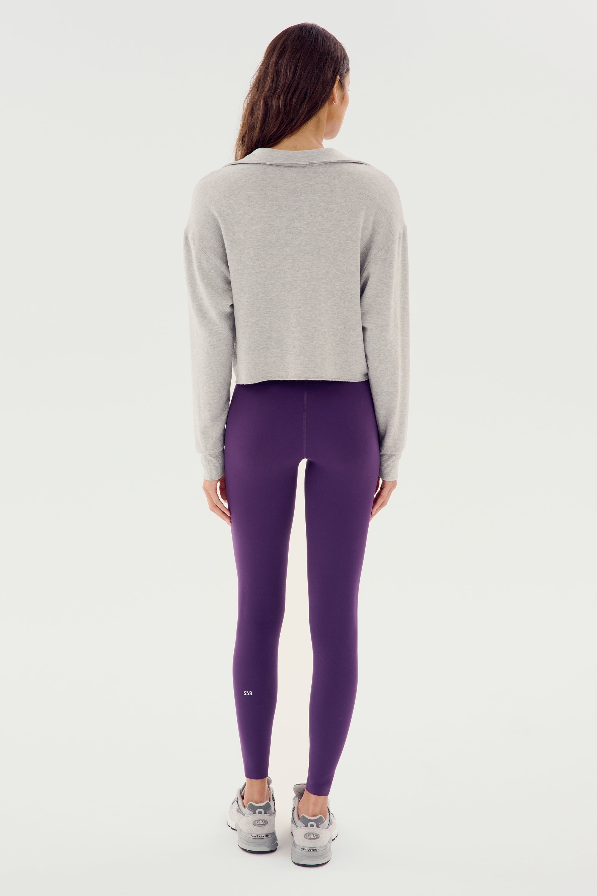 A woman is standing with her back to the camera, wearing a gray Joan Fleece Polo Sweatshirt by SPLITS59 made of 95% modal and 5% spandex, purple leggings, and gray sneakers.