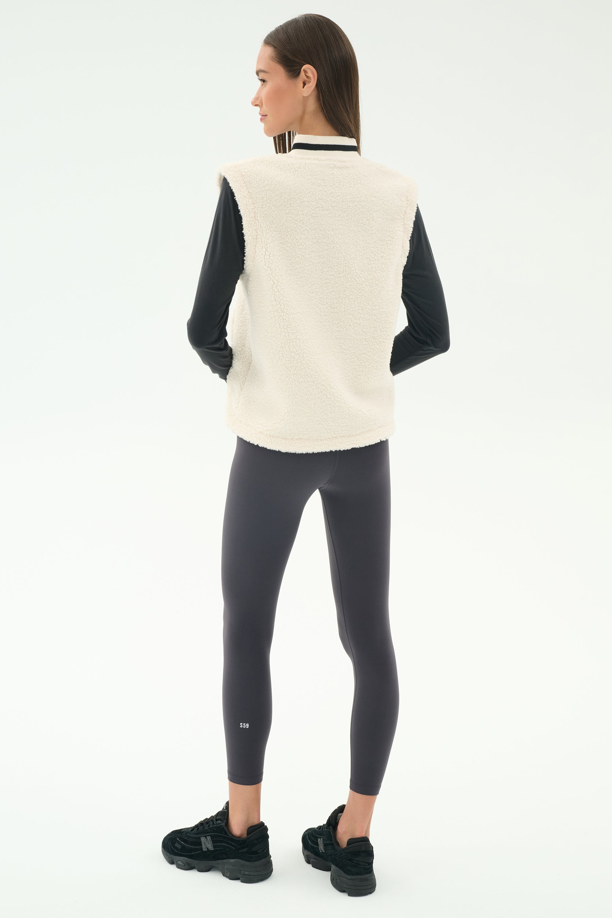 A woman faces away, wearing the SPLITS59 Aster Sherpa Vest in Creme over a black long-sleeve top, paired with dark gray leggings and black sneakers, set against a plain light background.