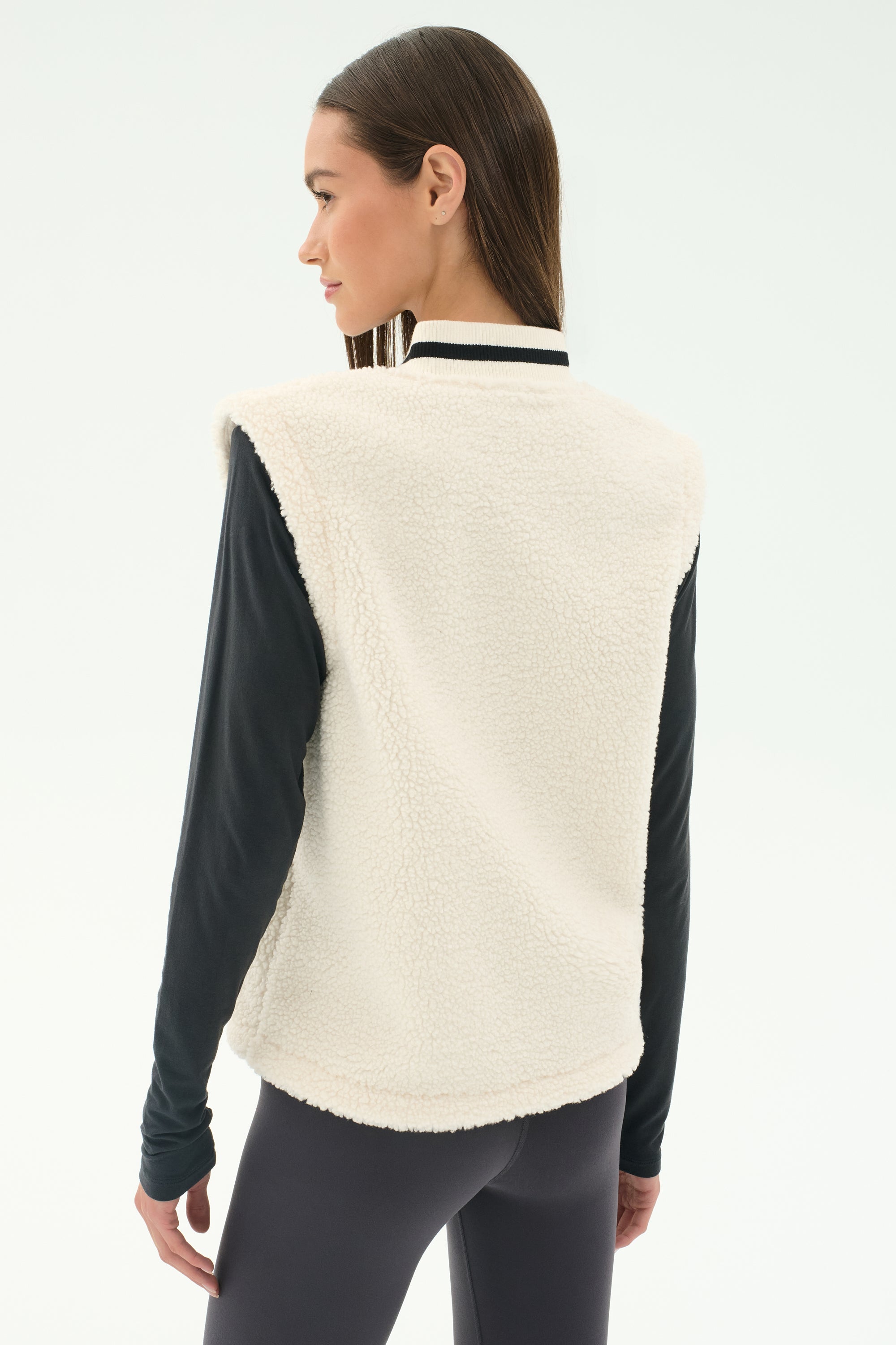 A woman stands with her back to the camera, wearing the SPLITS59 Aster Sherpa Vest in Creme over a black long-sleeve top and dark leggings.