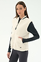 A woman wears the SPLITS59 Aster Sherpa Vest - Creme over a black long-sleeve top and leggings, standing against a plain white background, suited for cool weather workouts.
