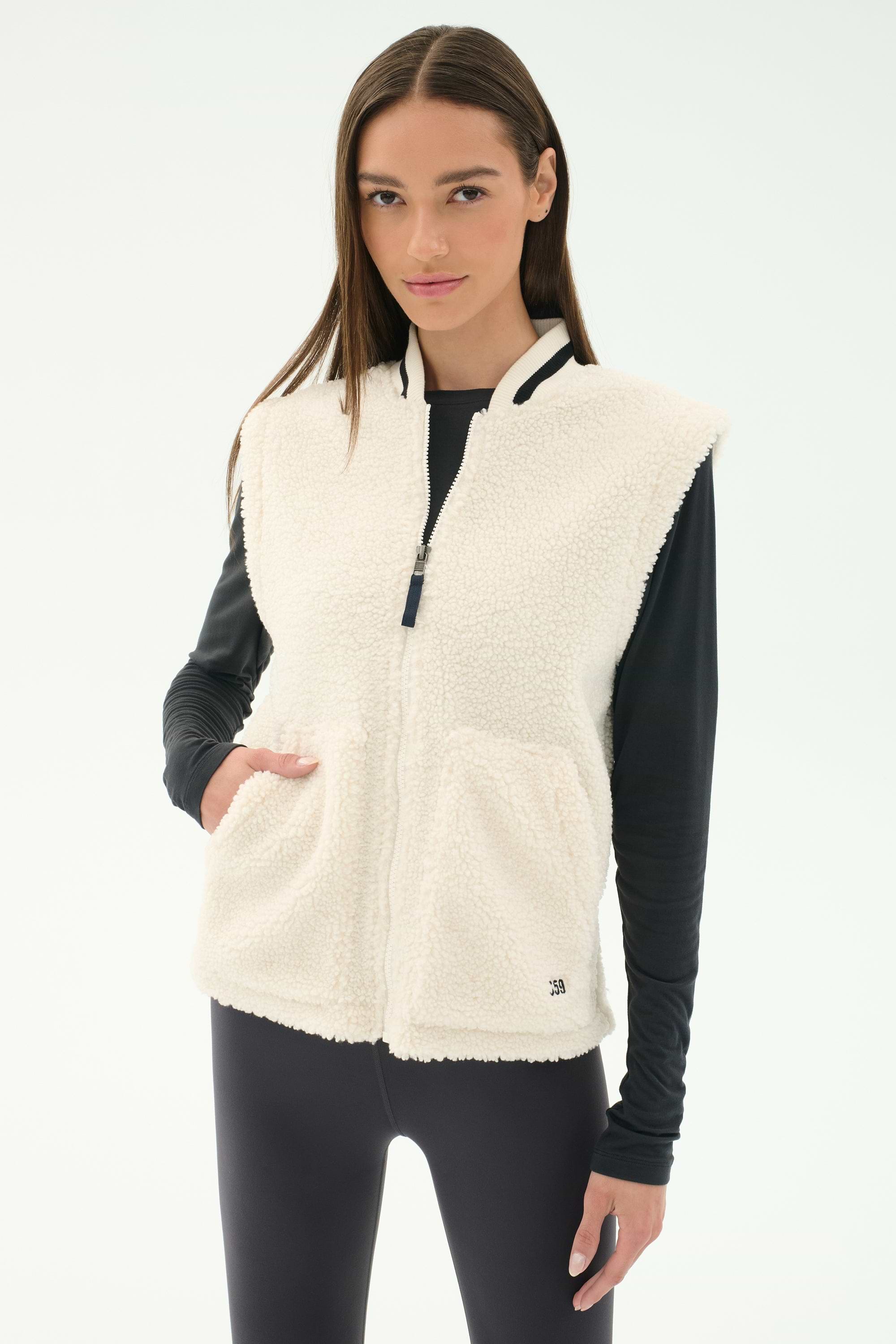 A woman stands against a plain light background wearing the SPLITS59 Aster Sherpa Vest - Creme over a black long-sleeve top and leggings—an ideal warm layer for cool weather workouts.