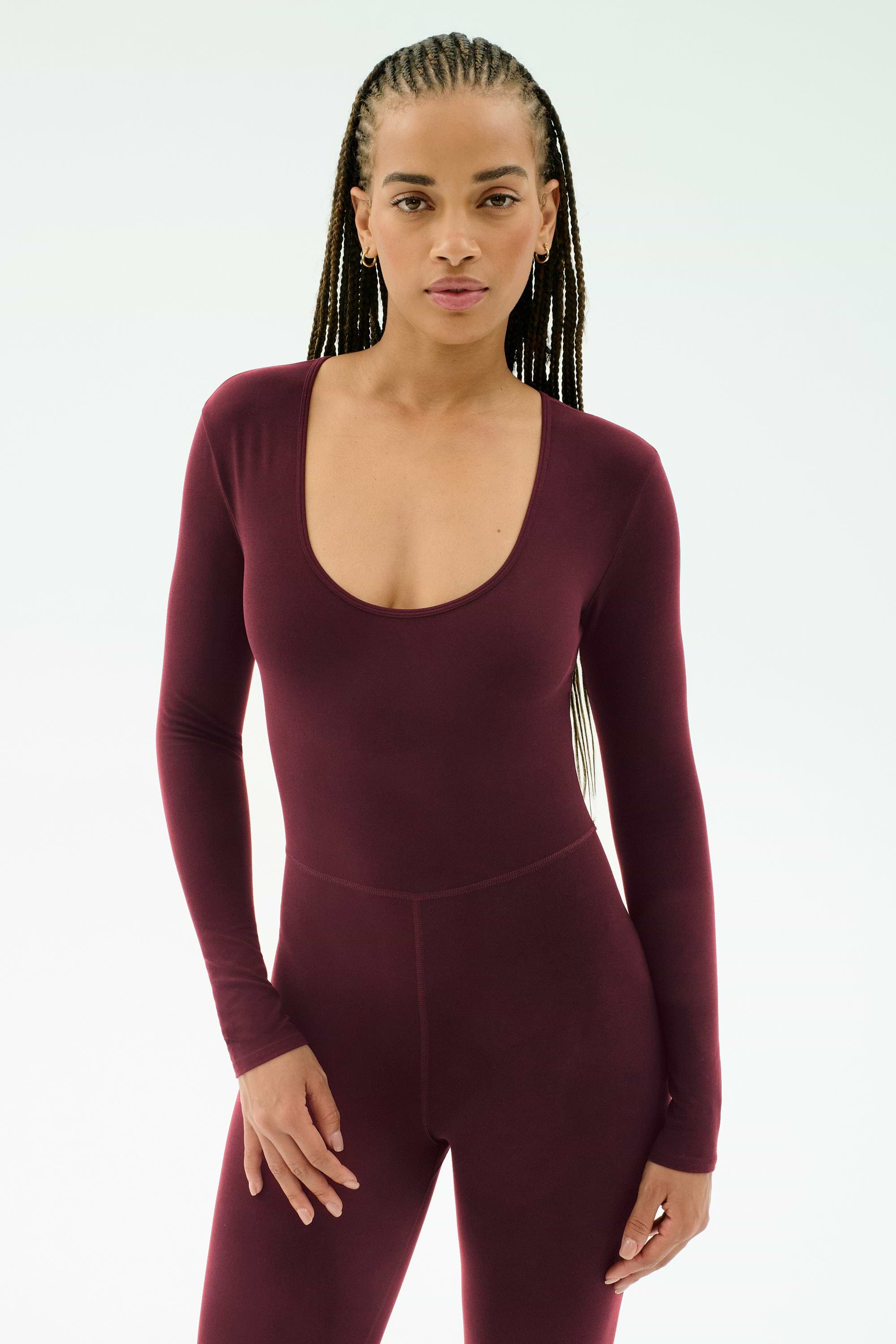 A woman with long braided hair wears the SPLITS59 Chloe Airweight Jumpsuit in Dark Cherry, featuring a fitted, long-sleeve design in a smooth nylon-spandex blend, standing against a plain light background.