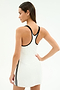 A woman in the SPLITS59 Ella Airweight Dress in White/Black, featuring a racerback and black trim, crafted from a nylon spandex blend, stands with hand on hip, facing away from the camera.