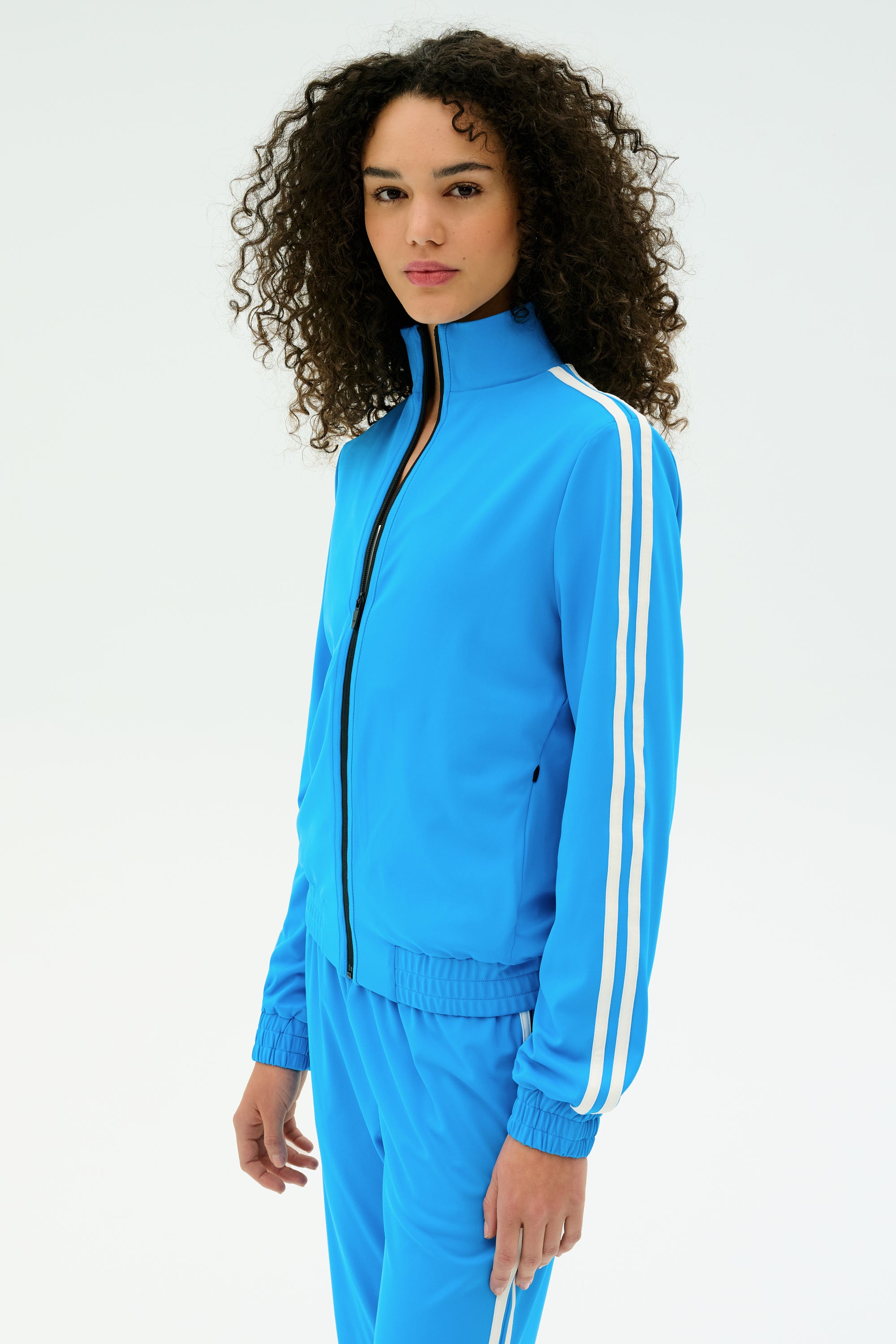 A person with curly hair wears the SPLITS59 Fox Techflex Jacket in Riviera/White, featuring a retro vibe and white sleeve stripes, standing against a plain white background.