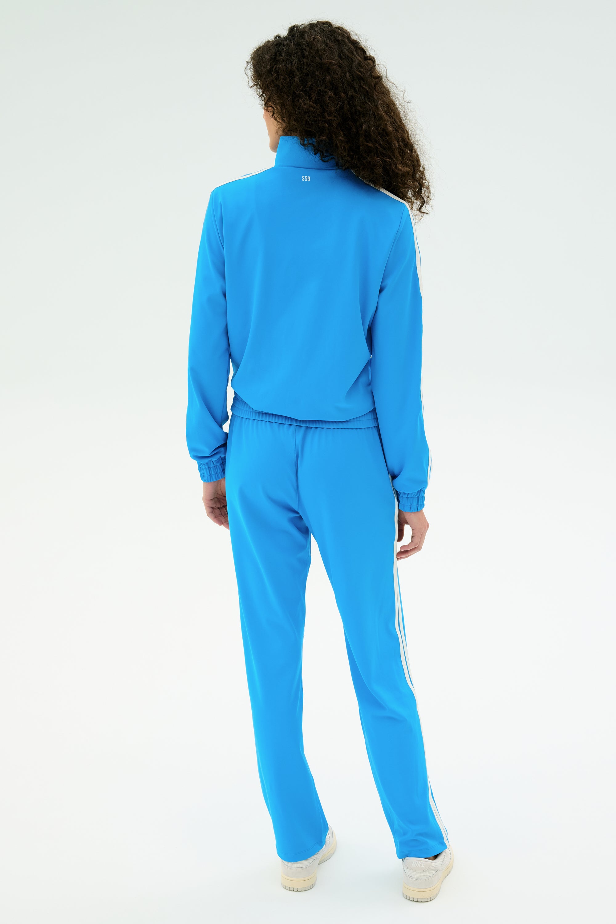 Person with curly hair wears the SPLITS59 Fox Techflex Jacket - Riviera/White and matching track pants with white sneakers, standing back to camera against a plain white background for a cool retro vibe.