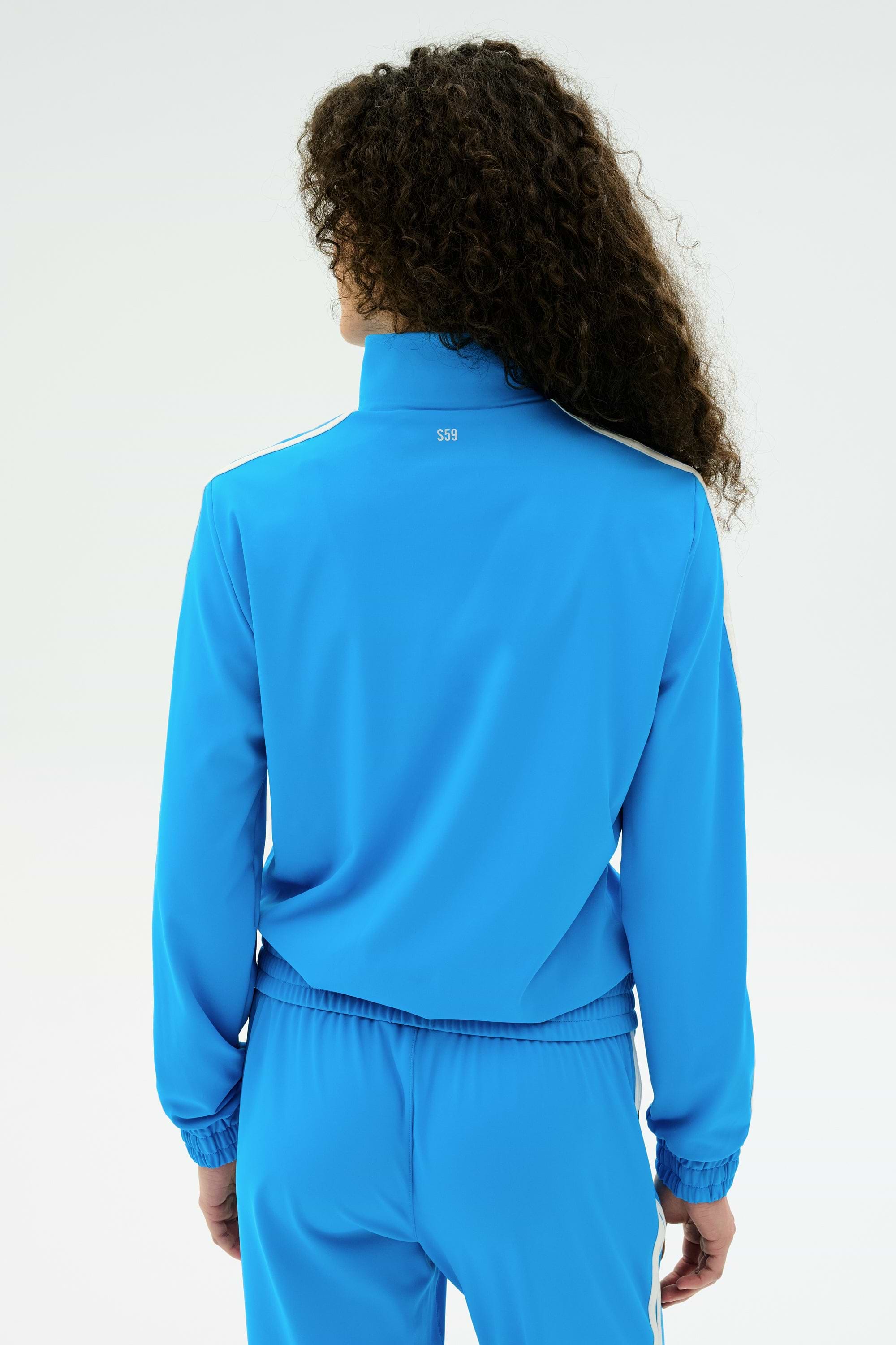 Person with long curly hair wears the SPLITS59 Fox Techflex Jacket in Riviera/White and matching Fox track pant, shown from the back against a plain light background for a retro vibe.