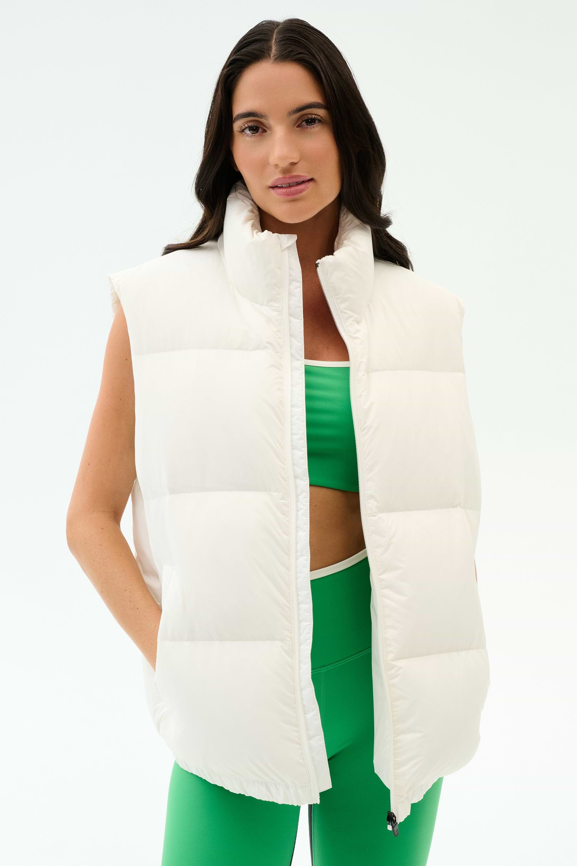 A woman models the SPLITS59 Summit Nylon Puffer Vest in white over a green sports bra and matching leggings, standing against a plain white background—ideal for lightweight insulation during cool weather workouts.