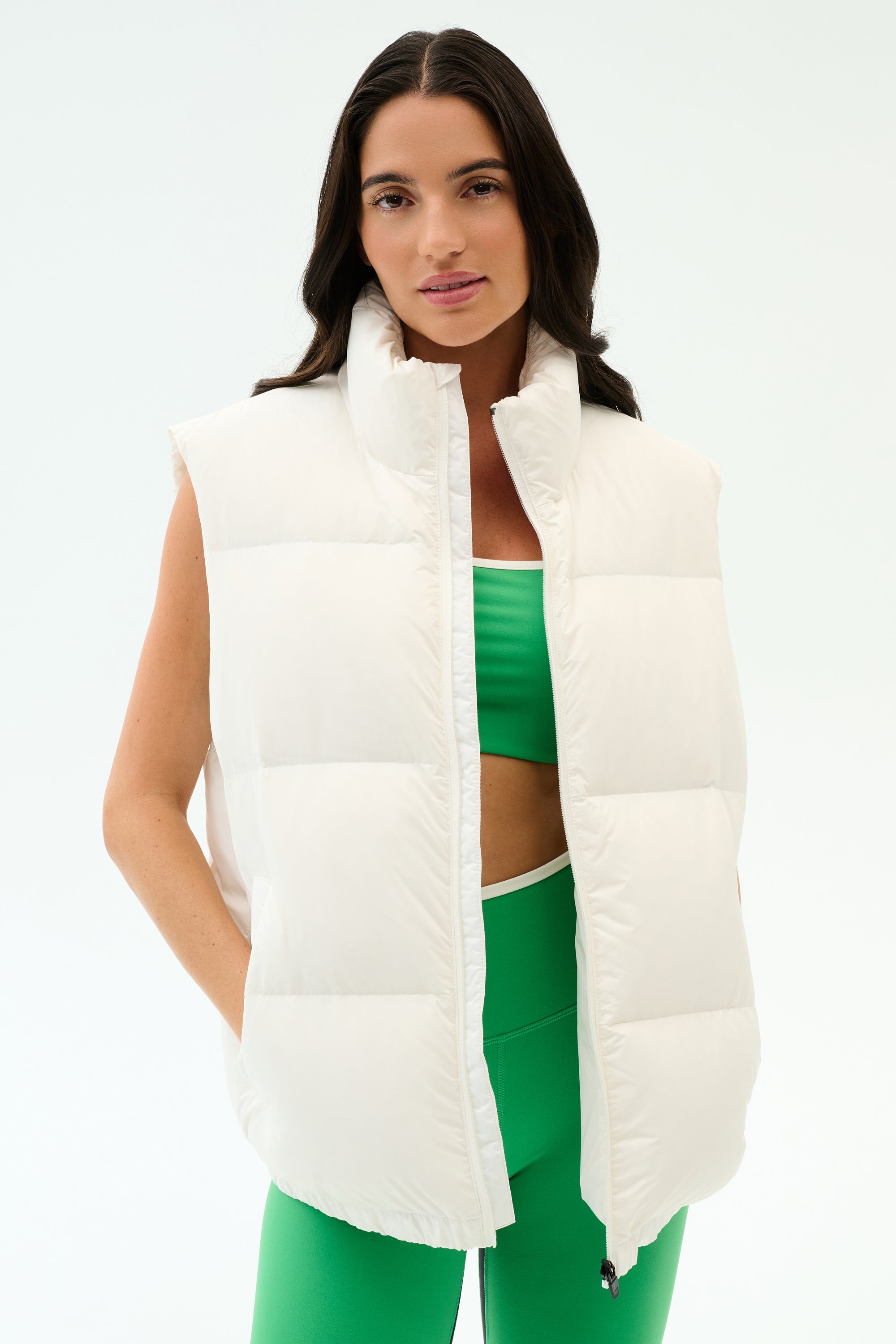 A woman models the SPLITS59 Summit Nylon Puffer Vest in white over a green sports bra and matching leggings, standing against a plain white background—ideal for lightweight insulation during cool weather workouts.