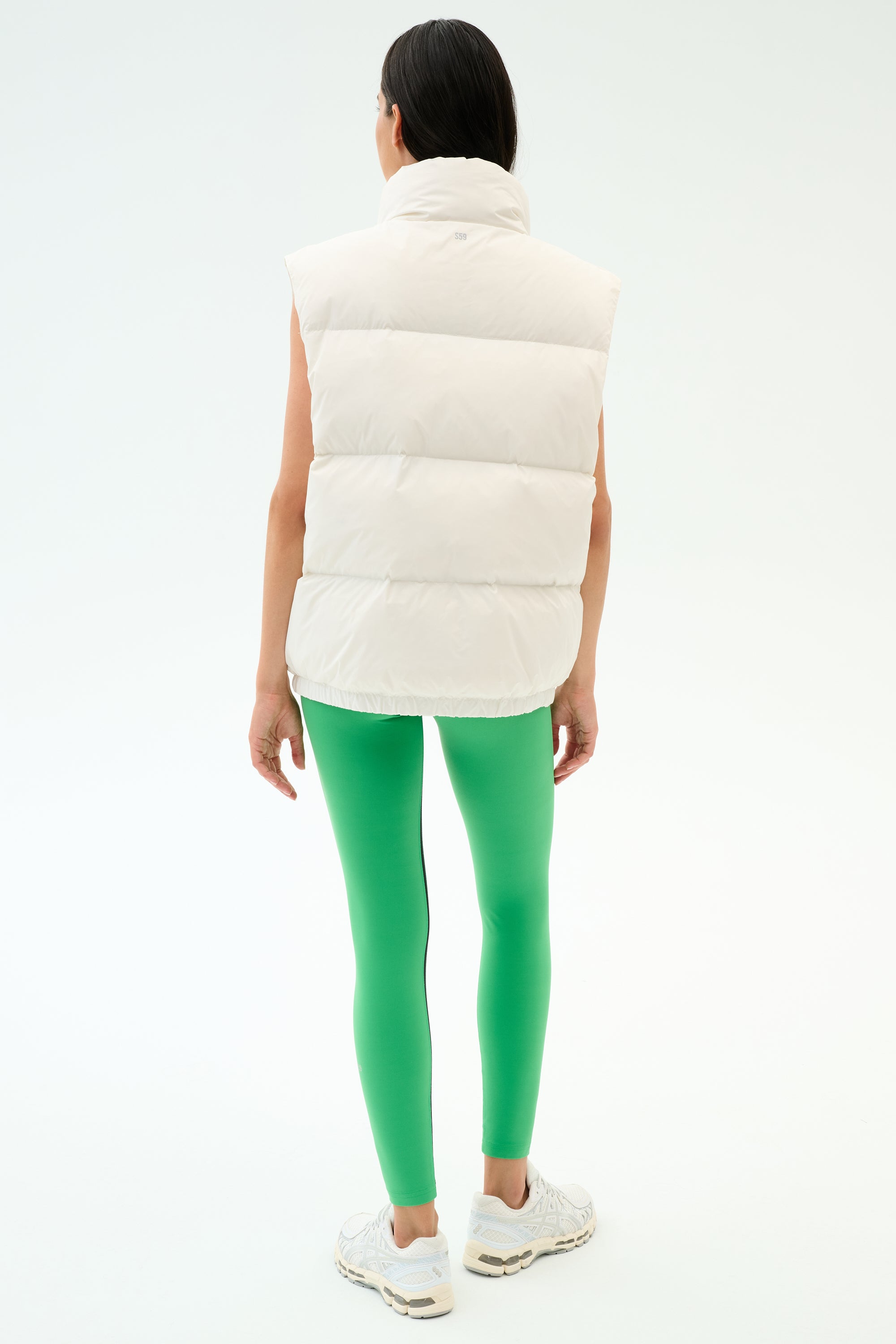 Wearing the SPLITS59 Summit Nylon Puffer Vest in white, paired with bright green leggings and white sneakers, a person stands facing away—showcasing a lightweight, stylish option for cool weather workouts against a plain background.
