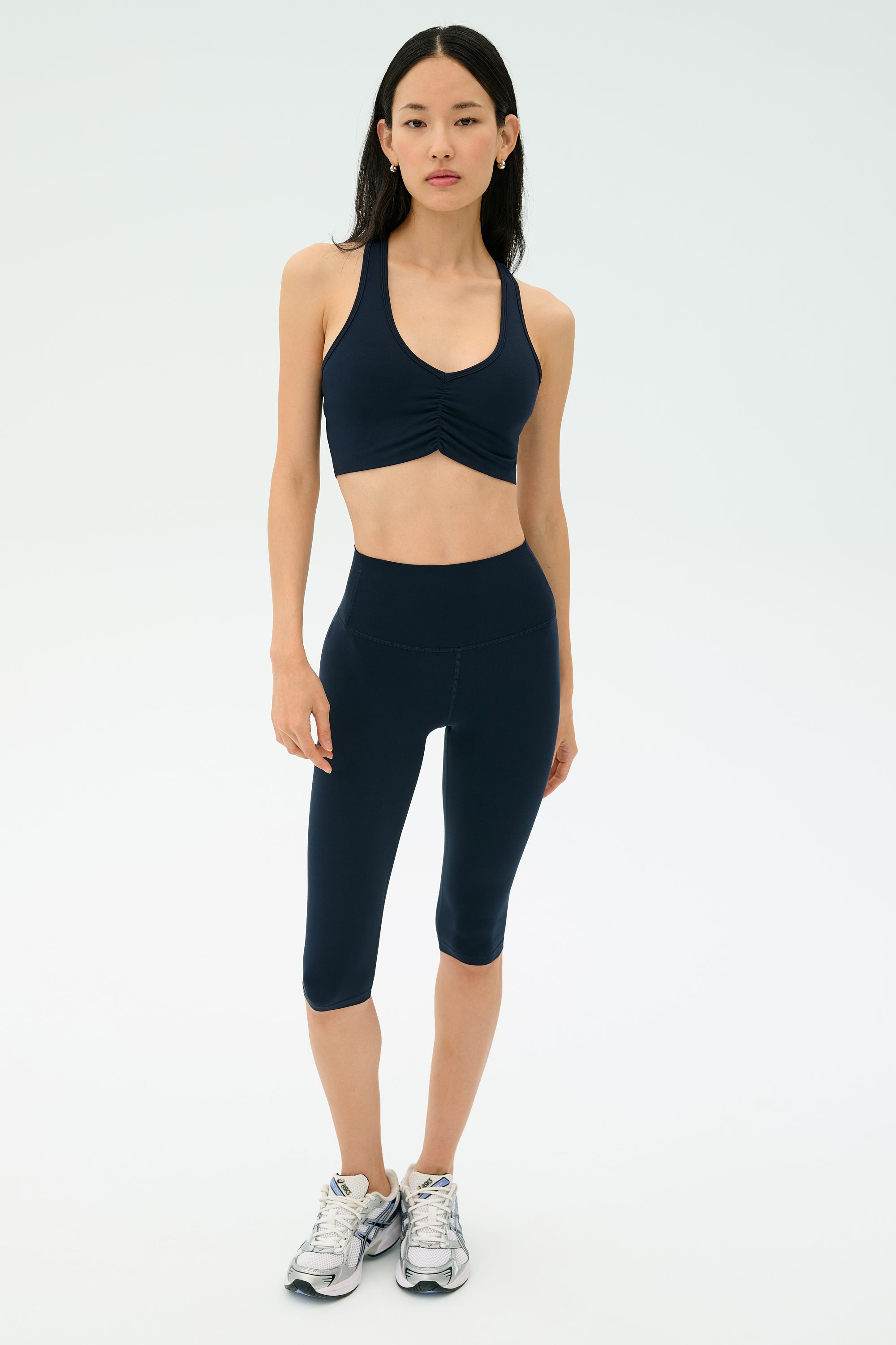 A woman stands facing forward against a white background, wearing the SPLITS59 Airweight High Waist 16" Legging in Indigo, a navy sports bra, and gray running shoes.
