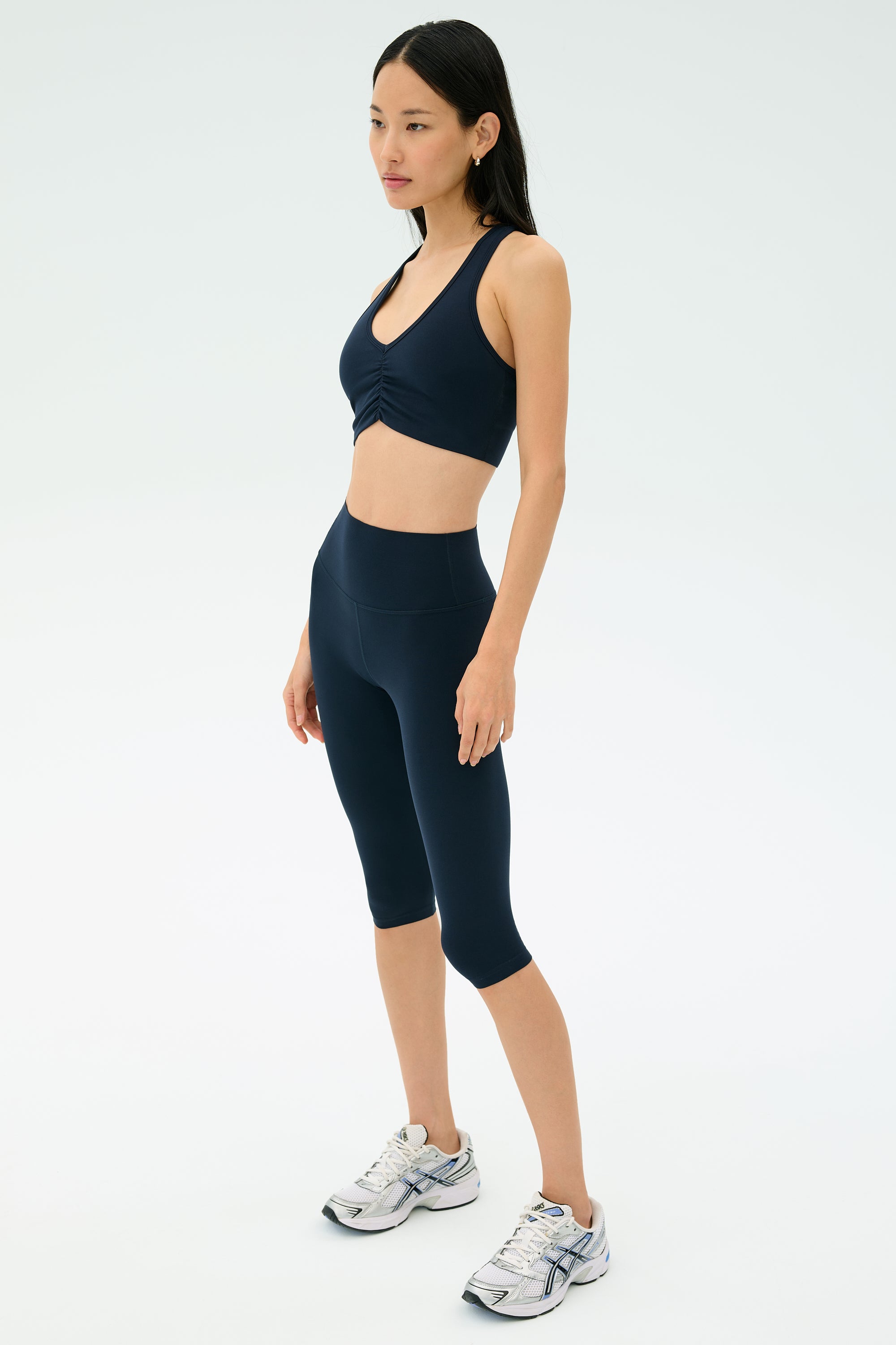 A woman stands in a studio wearing a navy sports bra, SPLITS59 Airweight High Waist 16" Legging in Indigo, and white athletic shoes.