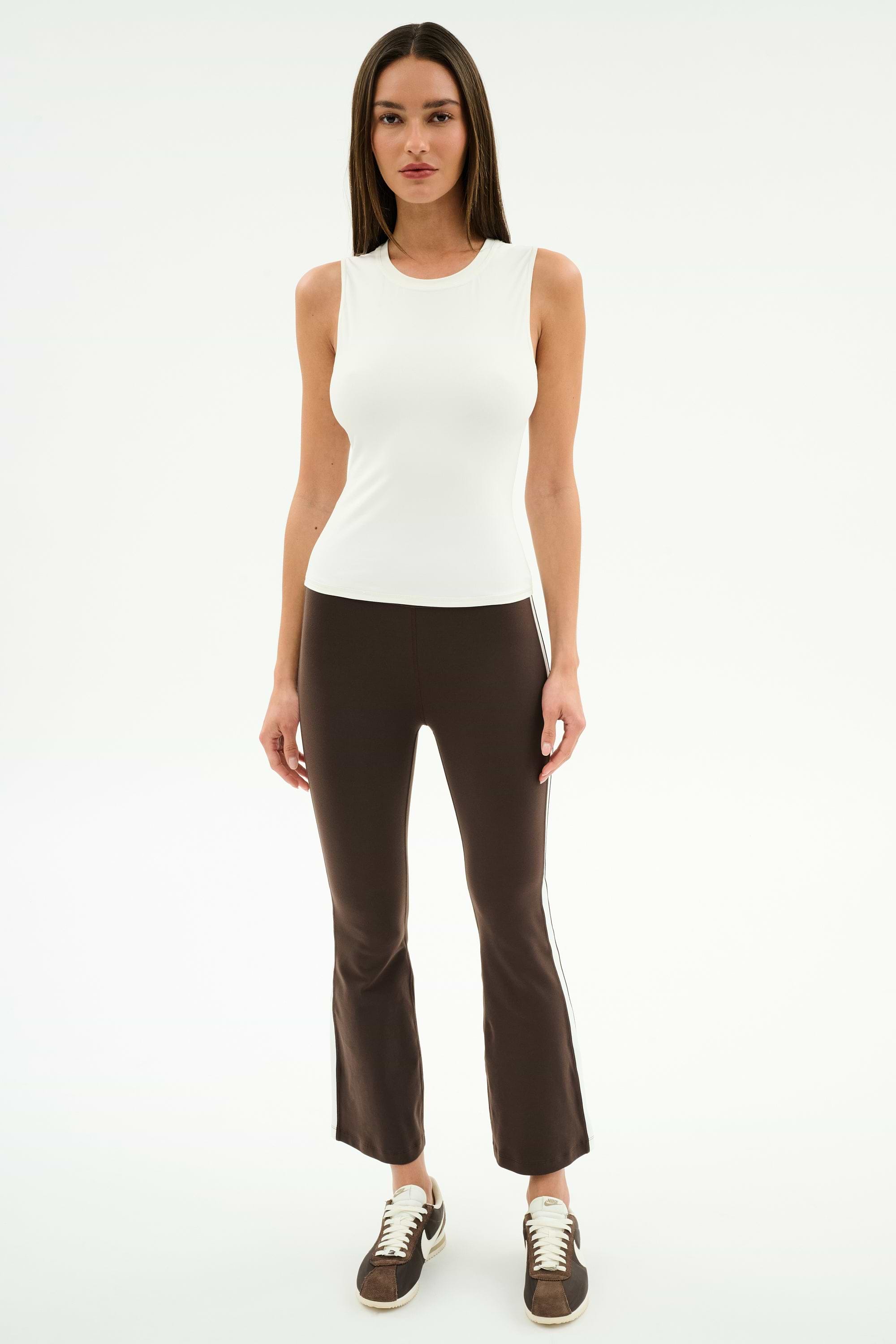 A woman faces forward against a white background, wearing the SPLITS59 Carter Airweight Lite Tank in white, paired with brown flared spandex pants with side stripes and brown sneakers.