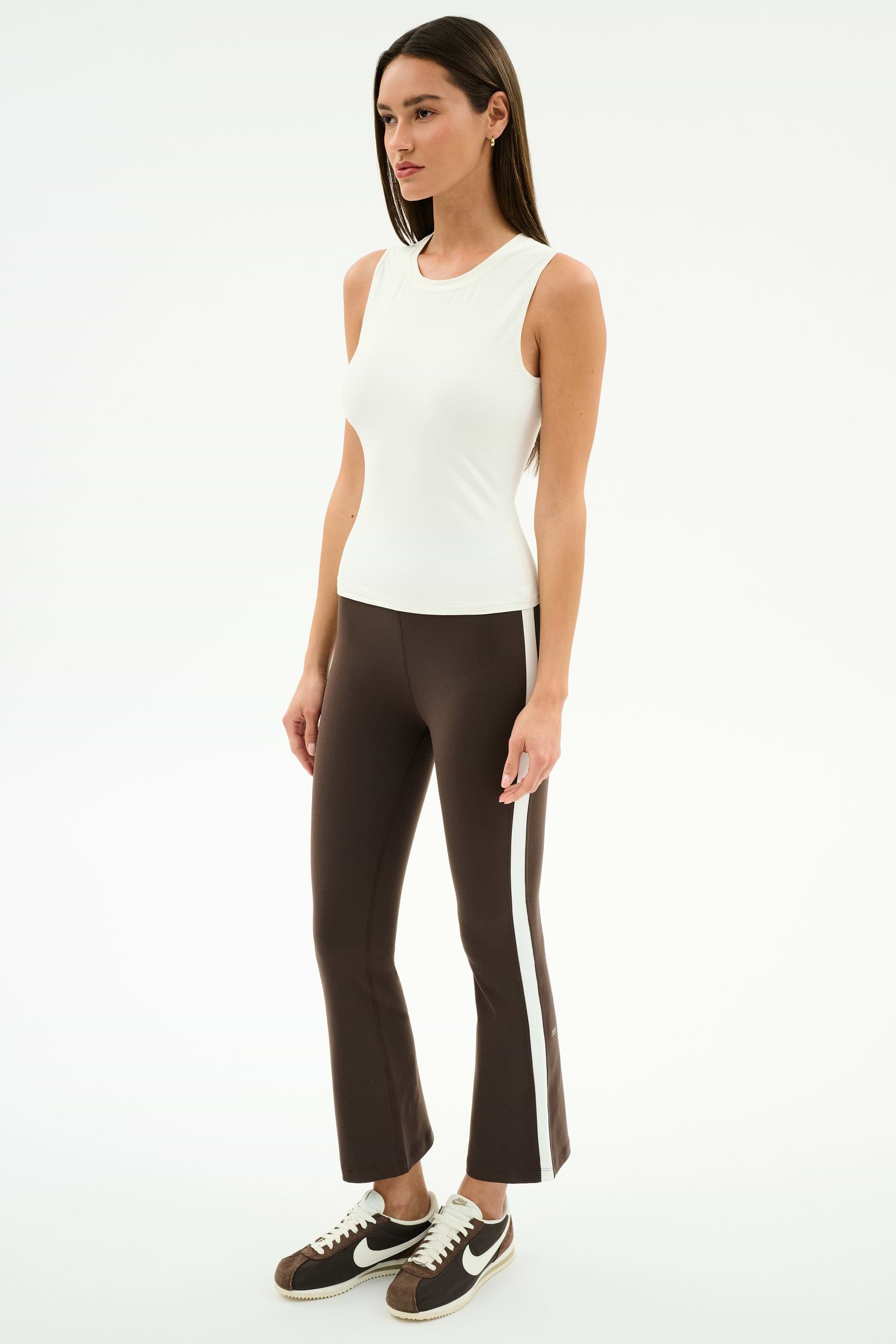 A woman stands against a plain white background wearing the SPLITS59 Carter Airweight Lite Tank in white, brown flared pants with a spandex side stripe, and brown Nike sneakers.