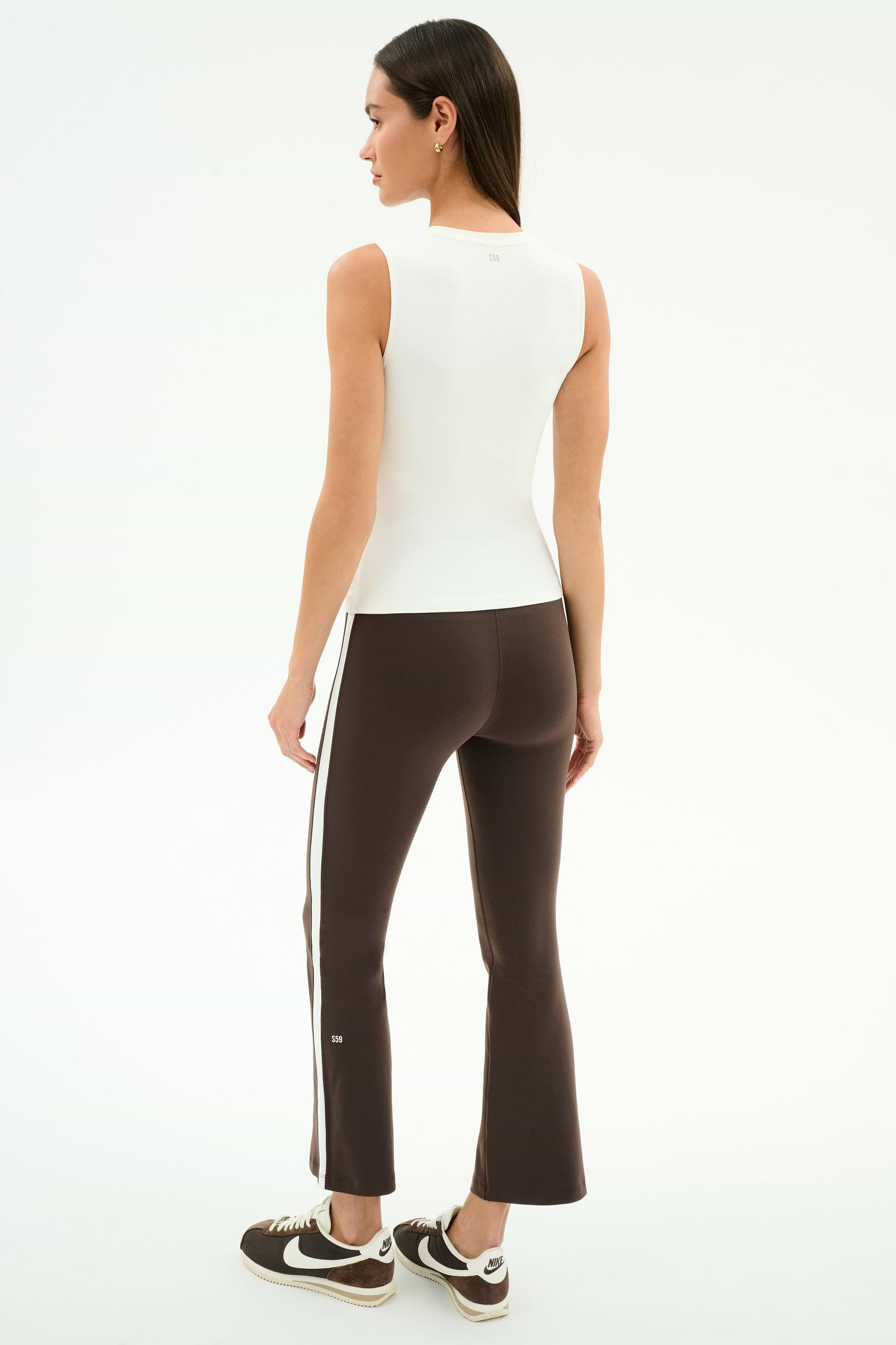 A woman faces away against a white background, wearing the SPLITS59 Carter Airweight Lite Tank in white, brown flared pants with side stripes, and brown sneakers.