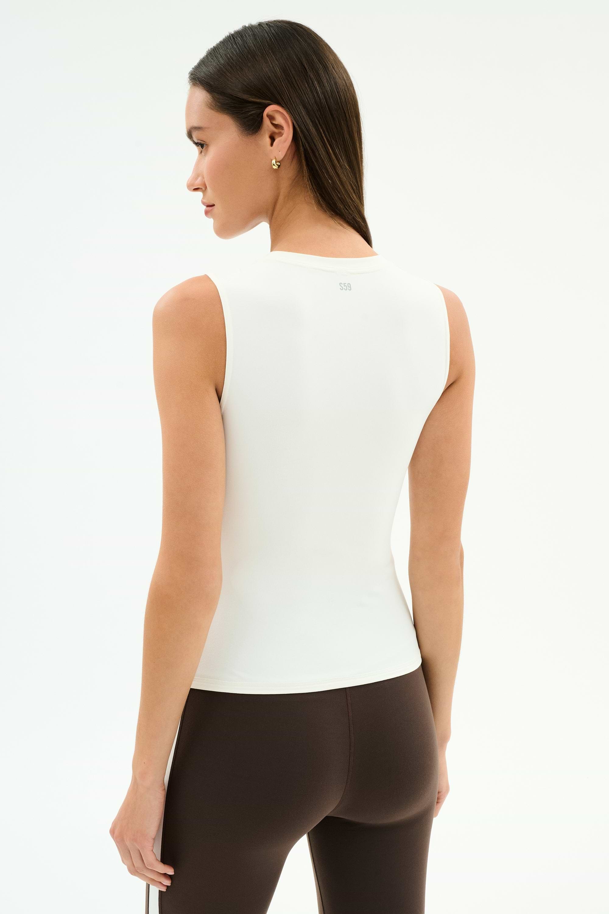 A woman faces away, wearing the SPLITS59 Carter Airweight Lite Tank in white and dark brown spandex leggings, set against a plain light background.