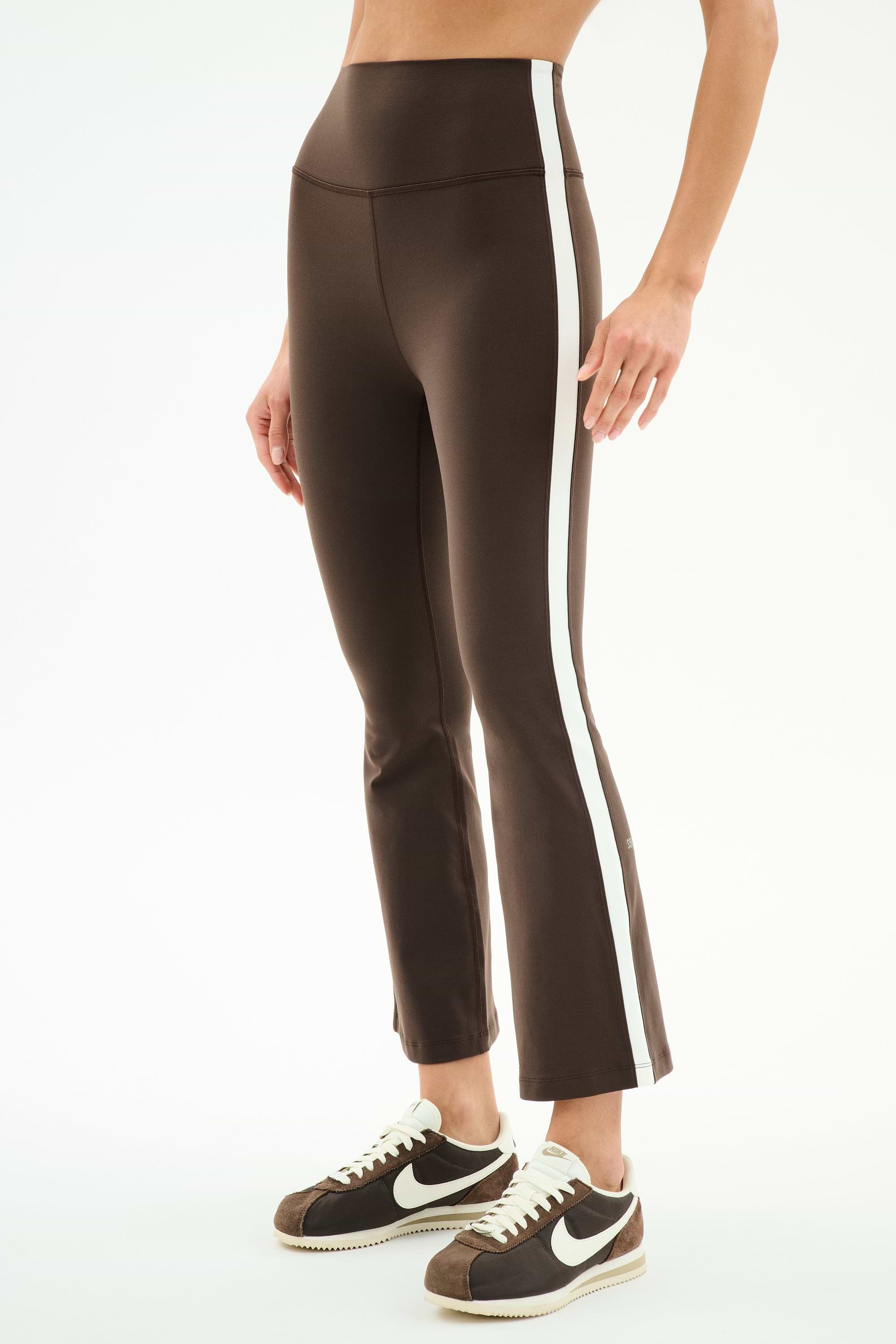 A person wears SPLITS59 Raquel High Waist Supplex Crop pants in Dark Chocolate/White with white side stripes and brown Nike sneakers, standing against a plain background.