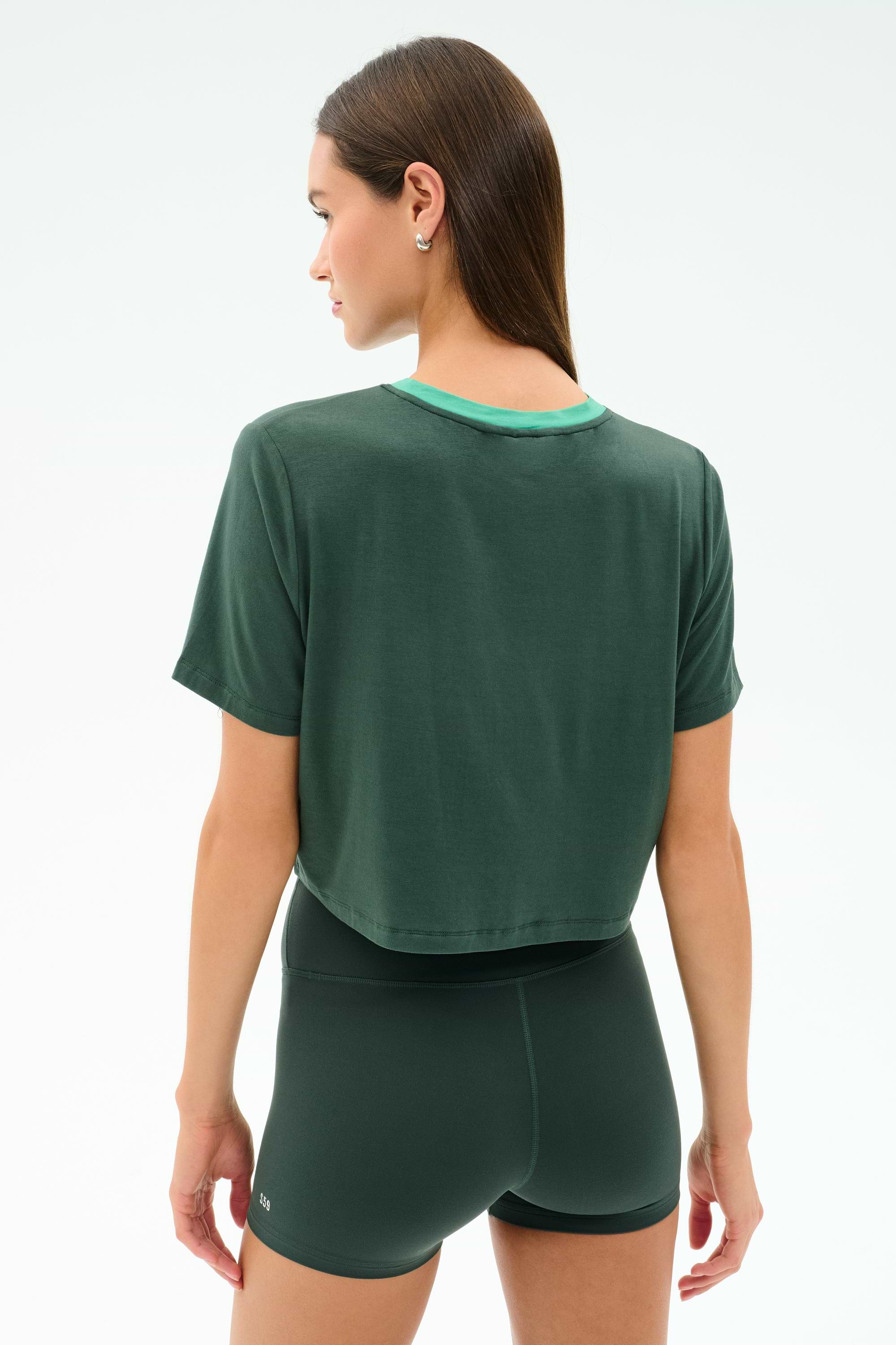 A person wearing the SPLITS59 Djuna Crop Ringer in Military/Spearmint, a dark green cropped t-shirt made from soft viscose with elastane for stretch, stands facing away against a plain light background.