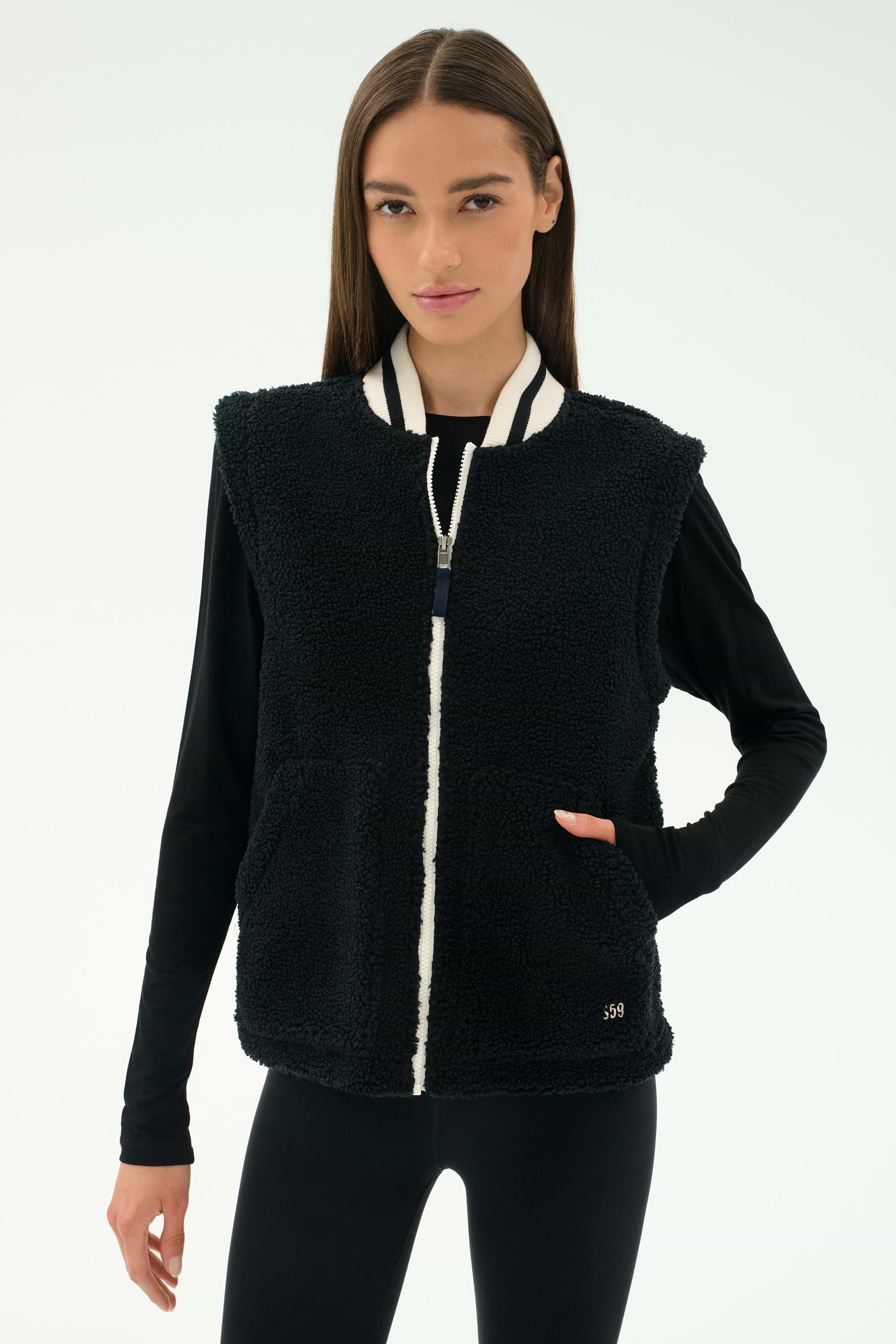 A woman stands against a plain light background, ready for cool weather workouts in the warm SPLITS59 Aster Sherpa Vest - Black, featuring a white zipper and striped collar over a long-sleeve black top and leggings.