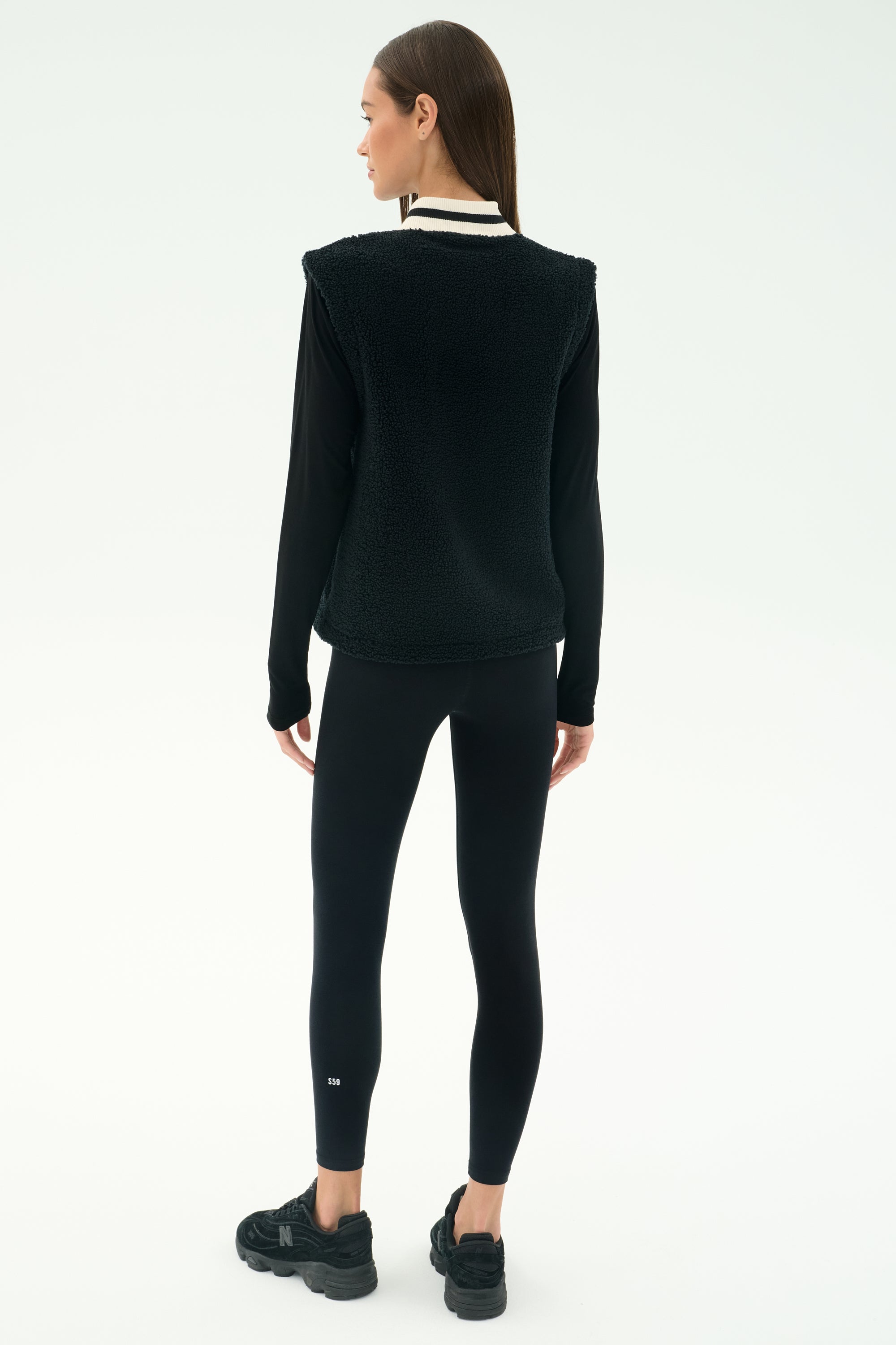 A woman is seen from behind wearing the SPLITS59 Aster Sherpa Vest in black over a long-sleeve top, paired with black leggings and sneakers, set against a simple light background.
