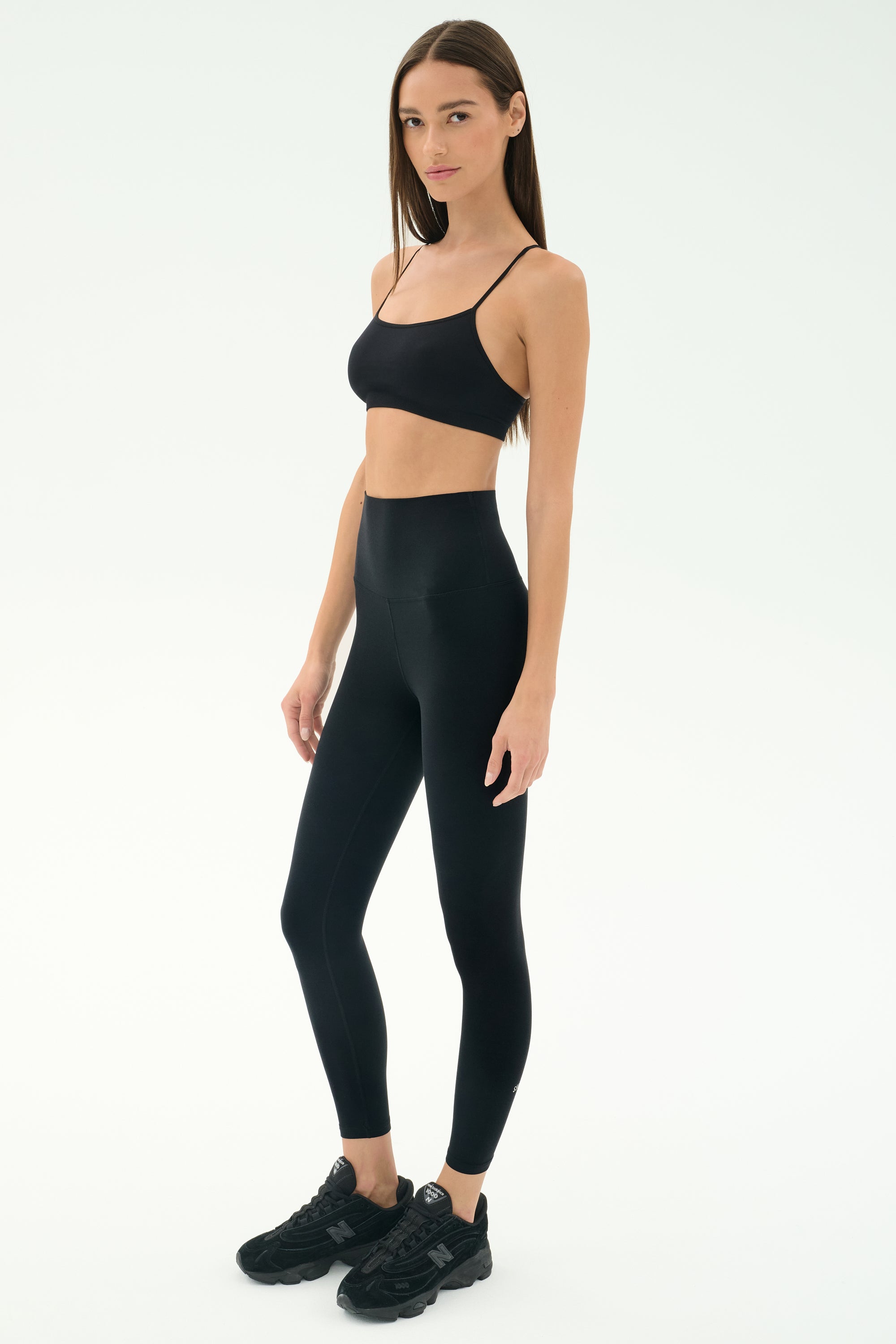 A woman stands against a white background wearing SPLITS59’s Airweight Super High Waist 7/8 leggings in black and a sports bra—breathable and ideal for hot yoga sessions.