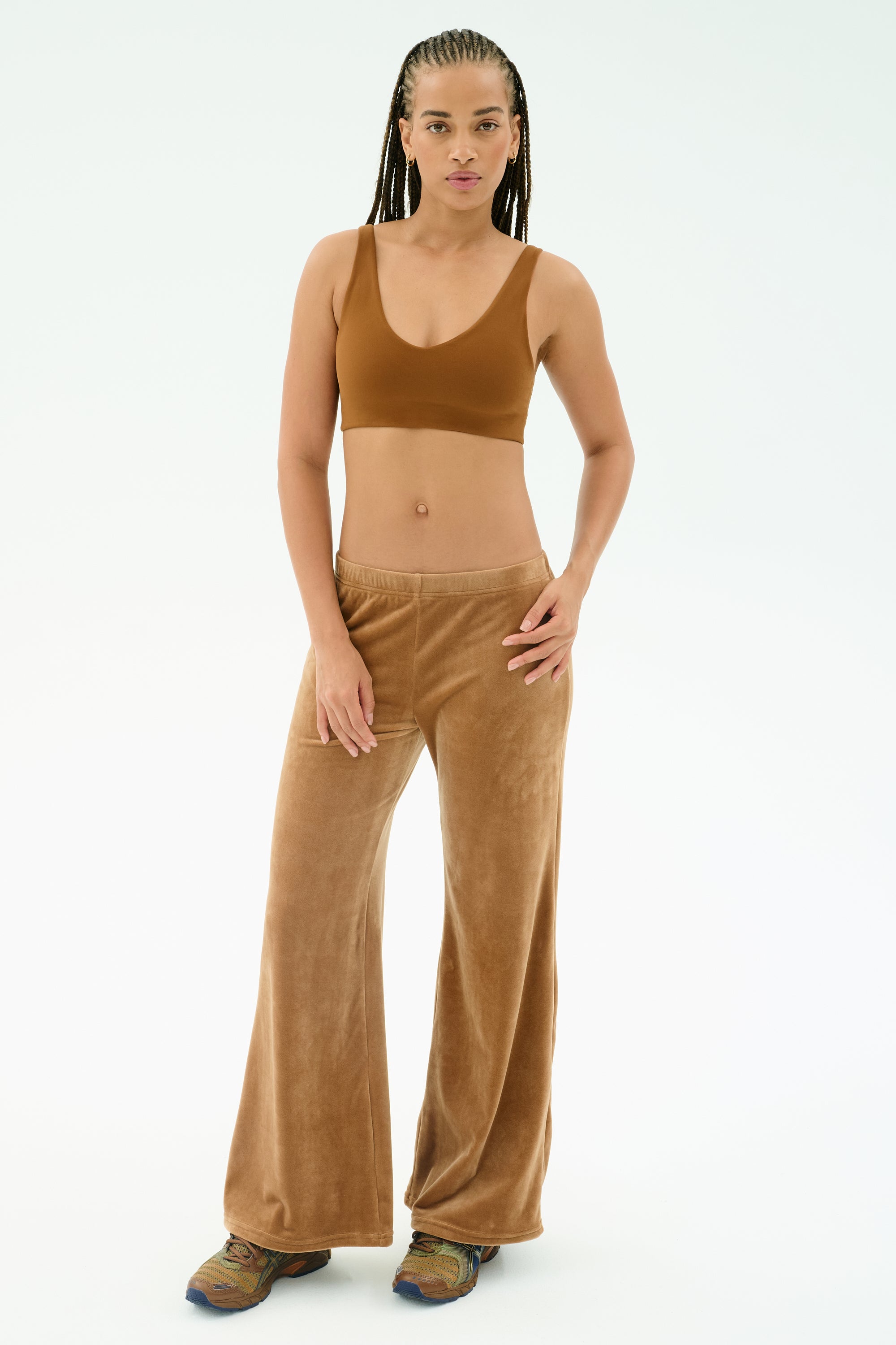 A person stands against a plain white background wearing SPLITS59’s Joy Velour Pant in Honey, crafted from soft polyester spandex fabric, paired with a matching brown crop top and colorful sneakers.