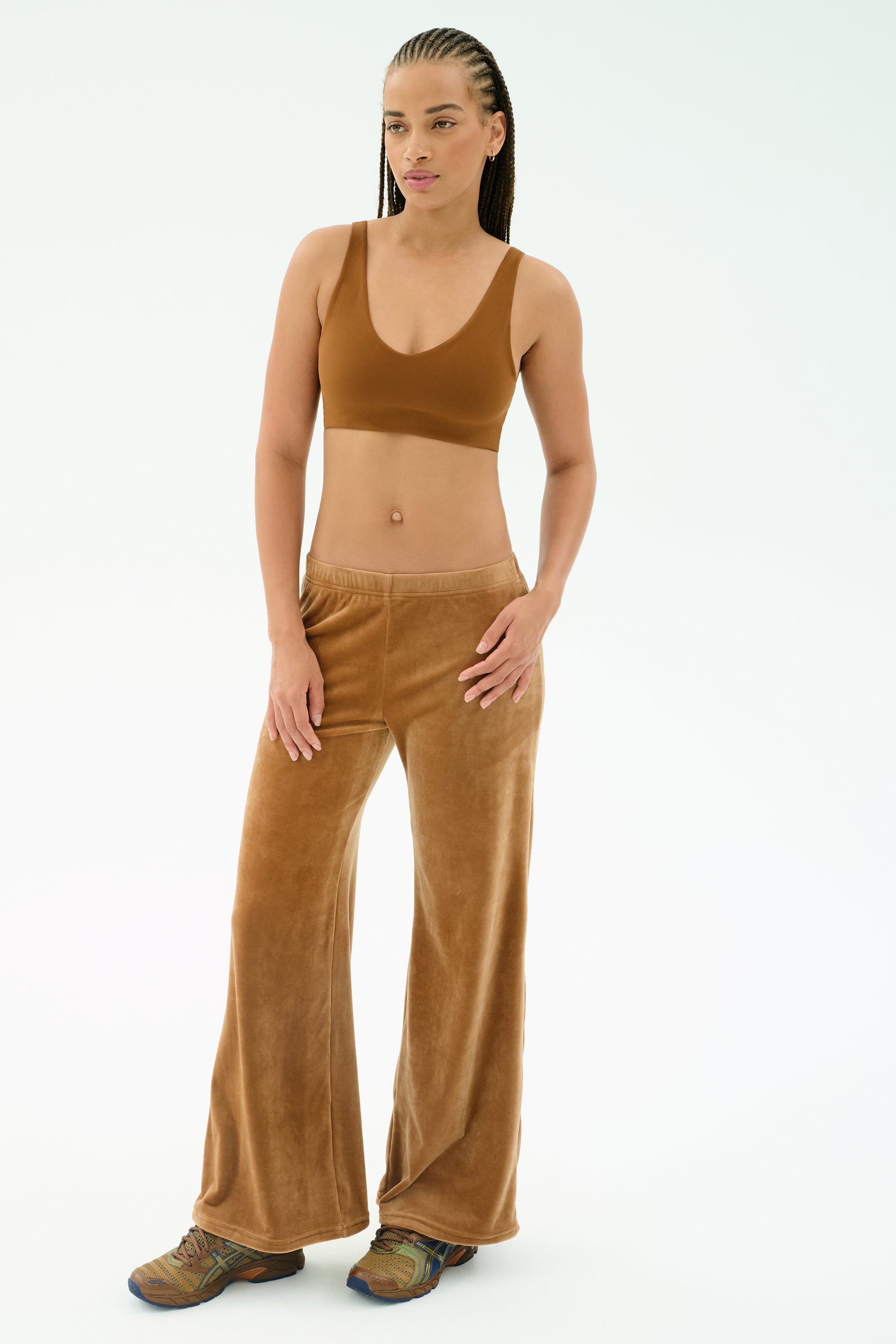 A woman stands against a plain background wearing the SPLITS59 Joy Velour Pant in Honey, a brown polyester spandex sports bra, and multicolored sneakers.